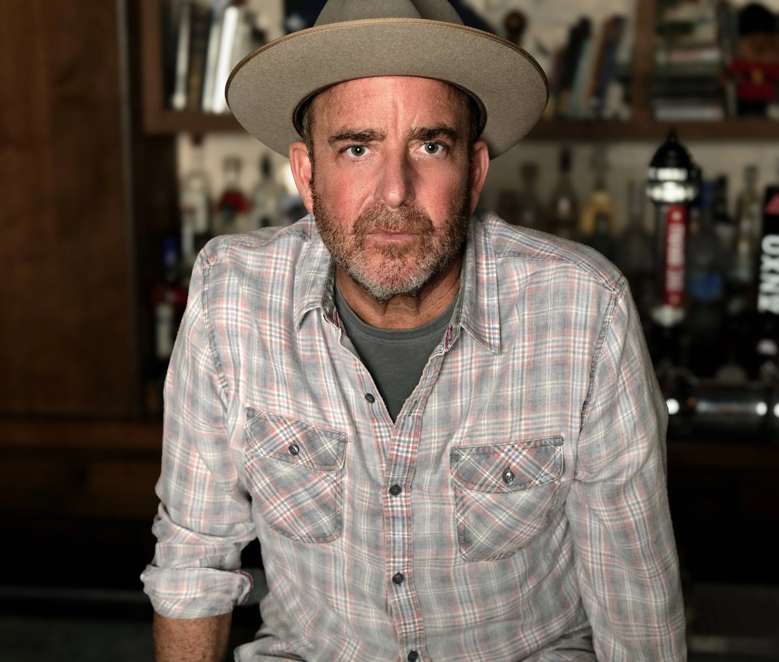 Close-up of a middle-aged man with a beard wearing a light-colored plaid shirt and a beige wide-brimmed hat, seated indoors with bookshelves and decorative items in the background.