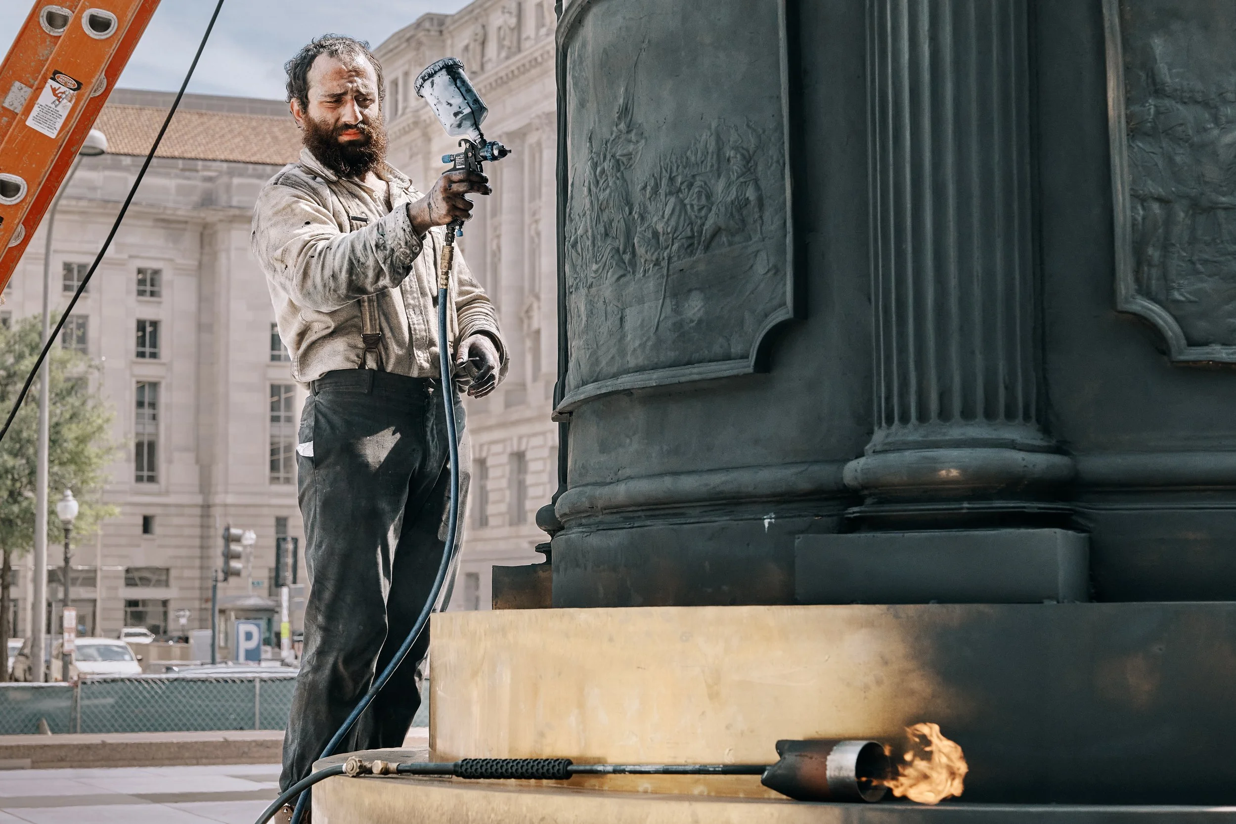 A man with a beard, wearing dirty clothes, adjusting a fire extinguisher near a large sculpture, with a flame coming out of a pipe at the bottom of the sculpture, in an urban setting.