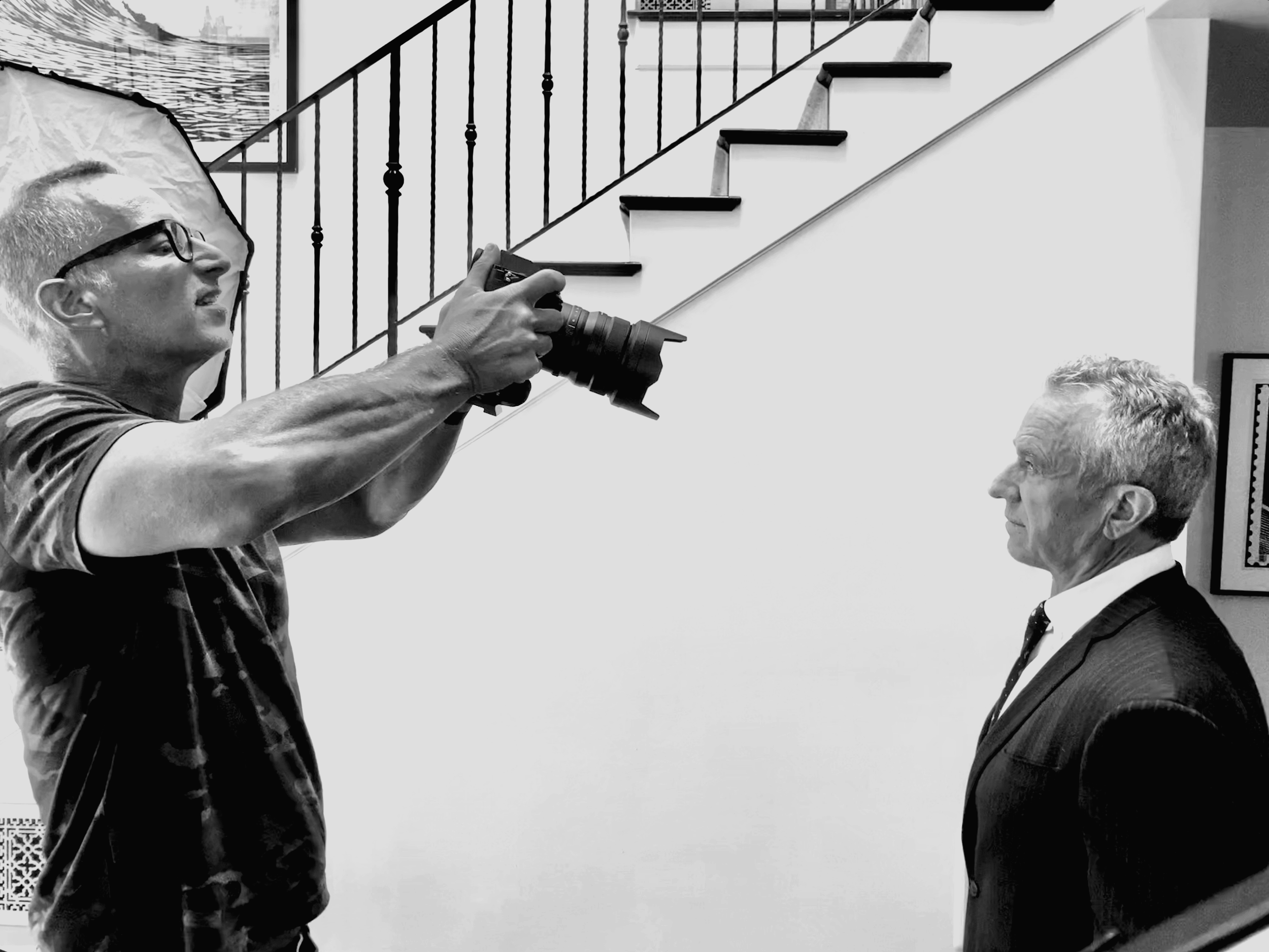 A man taking a photograph of an older man dressed in a suit, both indoors with a staircase and framed pictures on the wall in the background.