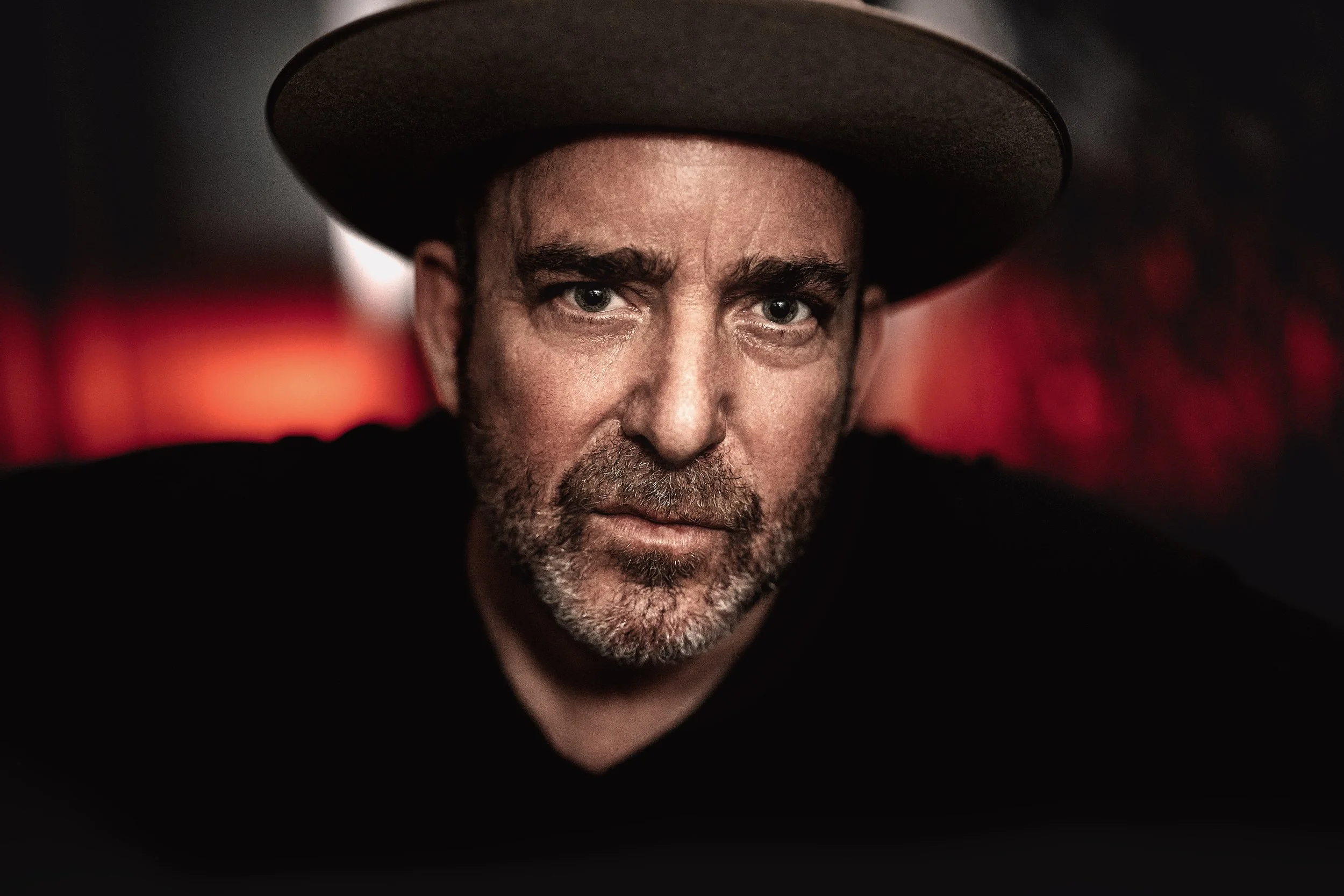 Close-up of a man, Jon Kahn, face with piercing blue eyes, gray beard, and dark hair underneath a wide-brimmed black hat, against a dark background with blurred red lights.