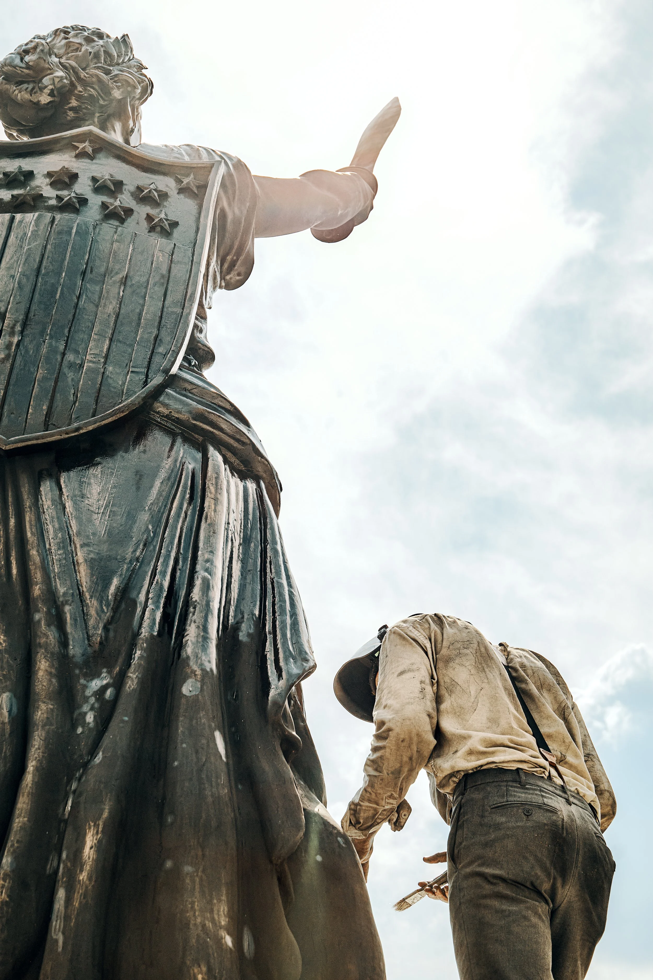 View from below of a sculptor standing next to a large bronze statue of a historical figure with a shield and a sword, sky with clouds in the background. Freedom Plaza in Washington, DC. 