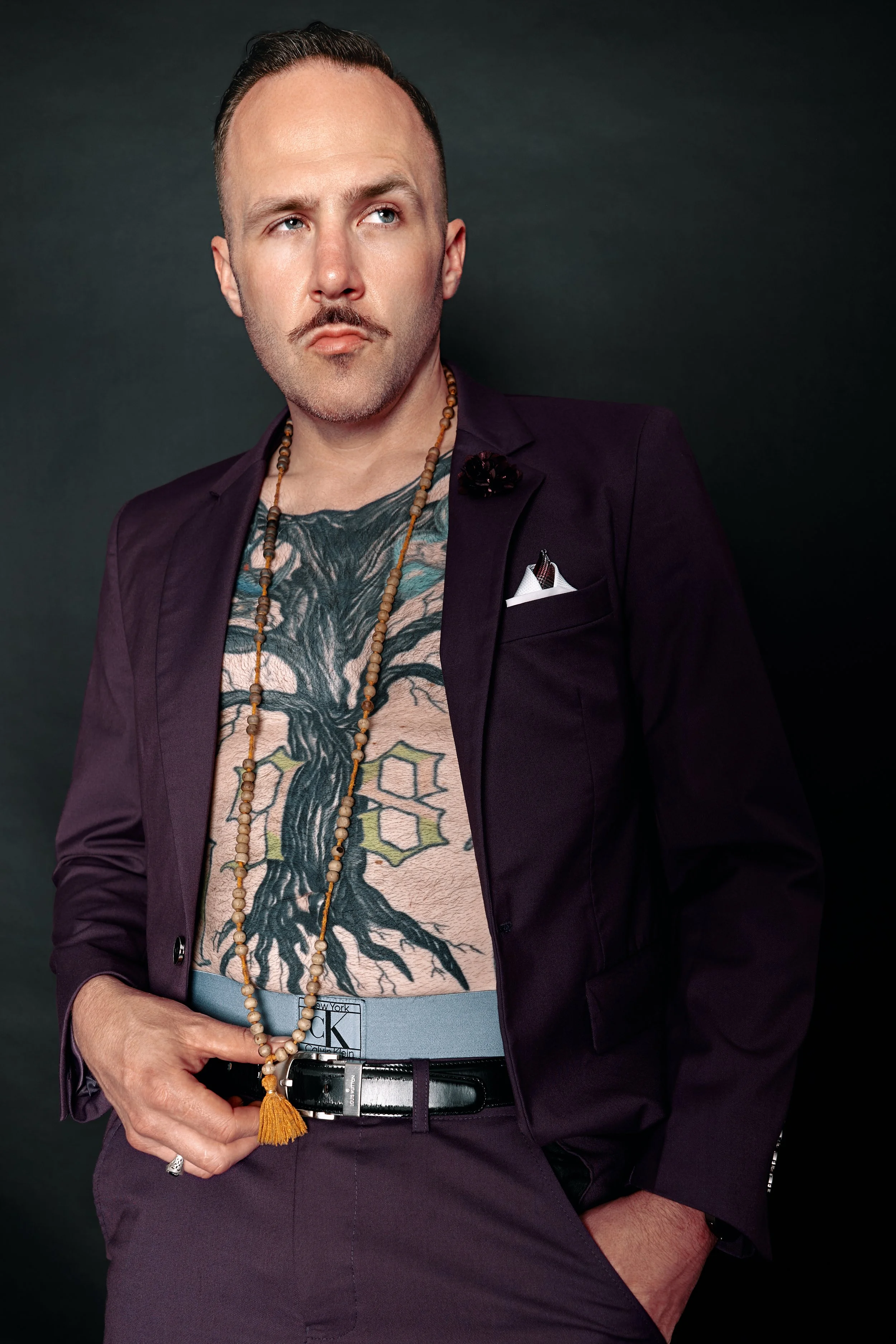 Portrait of a man with a mustache and beard, wearing a dark purple blazer, with tattoos on his chest showing a tree and hexagon patterns, accessorized with beaded necklaces, a pocket square, and a belt, against a dark background.