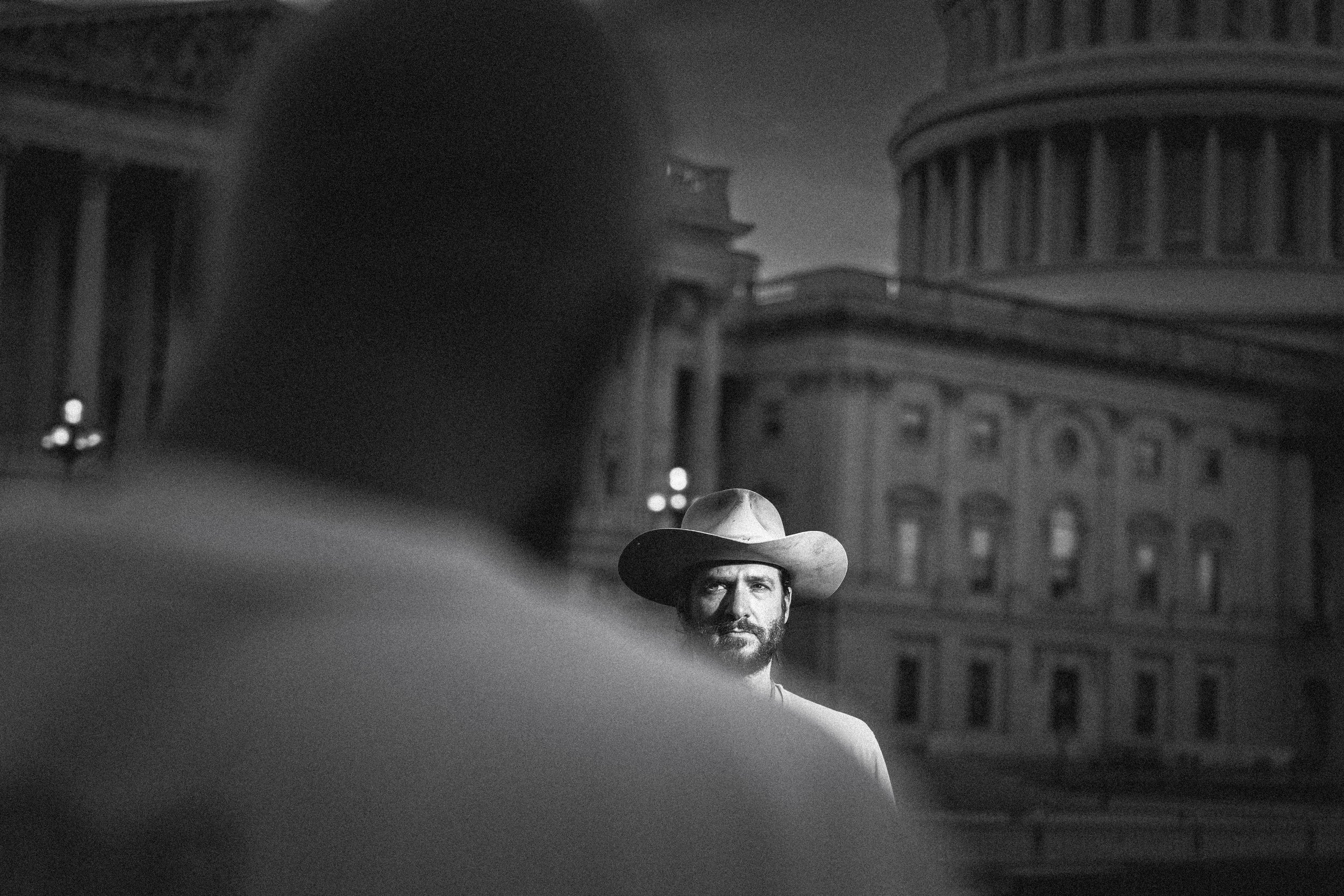A man wearing a cowboy hat gazes in the direction of the camera, partially obscured by a person in front of him, during a nighttime outdoor scene with historic buildings in the background.