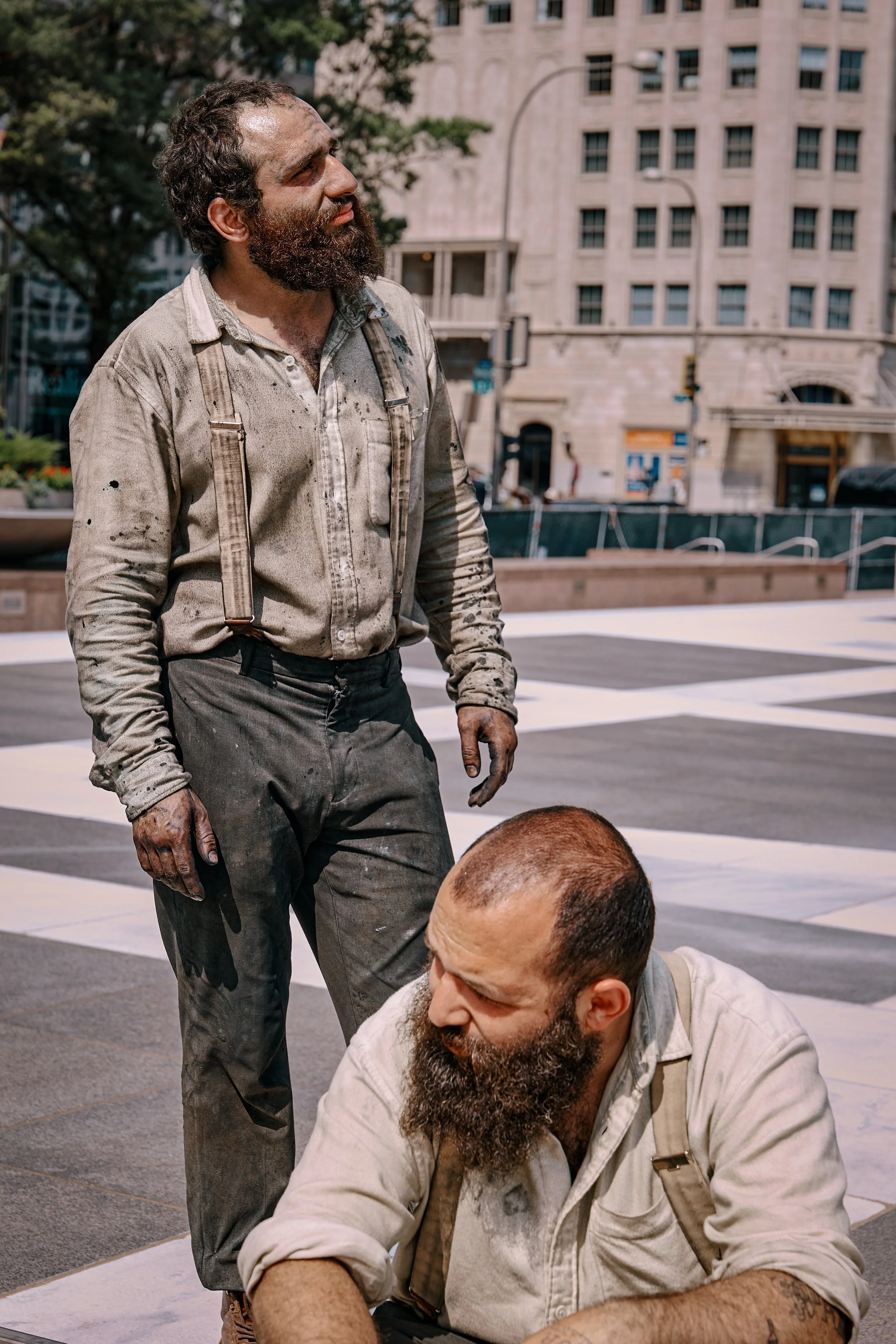 Two men with beards and dirty clothing sitting and standing on a city street, with a large building and trees in the background.
