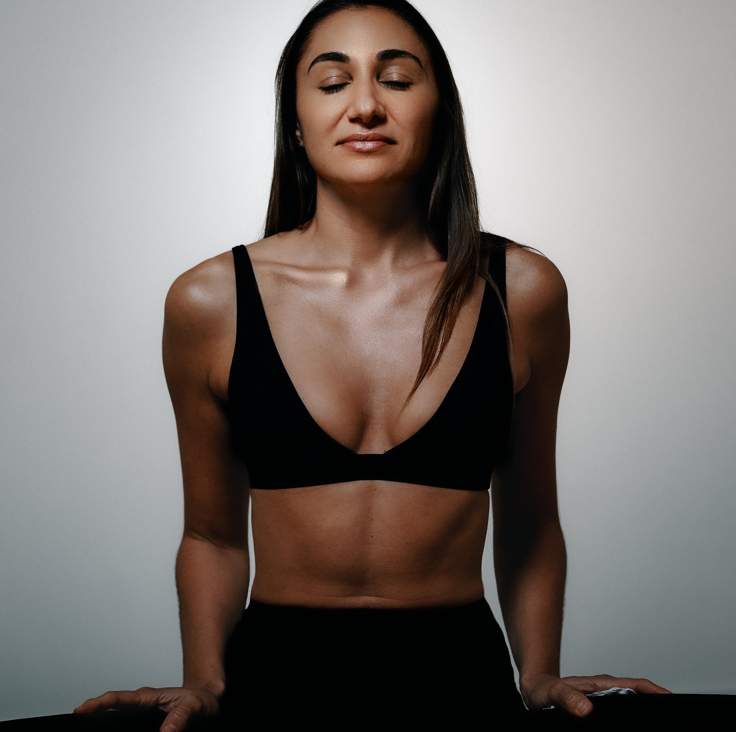 A beautiful woman with long dark hair and medium skin tone, wearing a black tank top, in a meditative posev with her eyes closed, and her hands resting on her legs. The background is plain and gray.