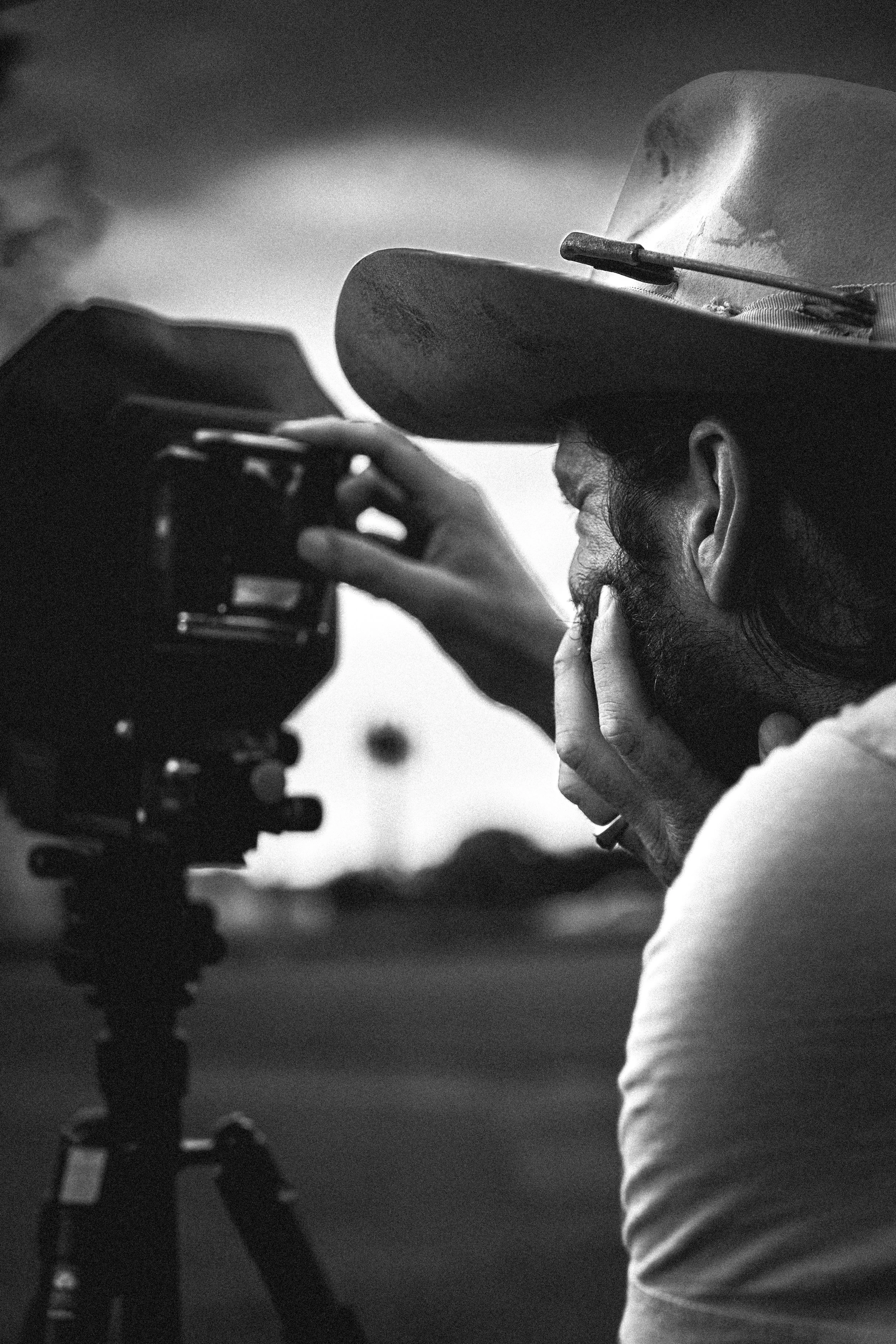 A person wearing a wide-brimmed hat looking through a camera on a tripod, possibly taking photos or filming.