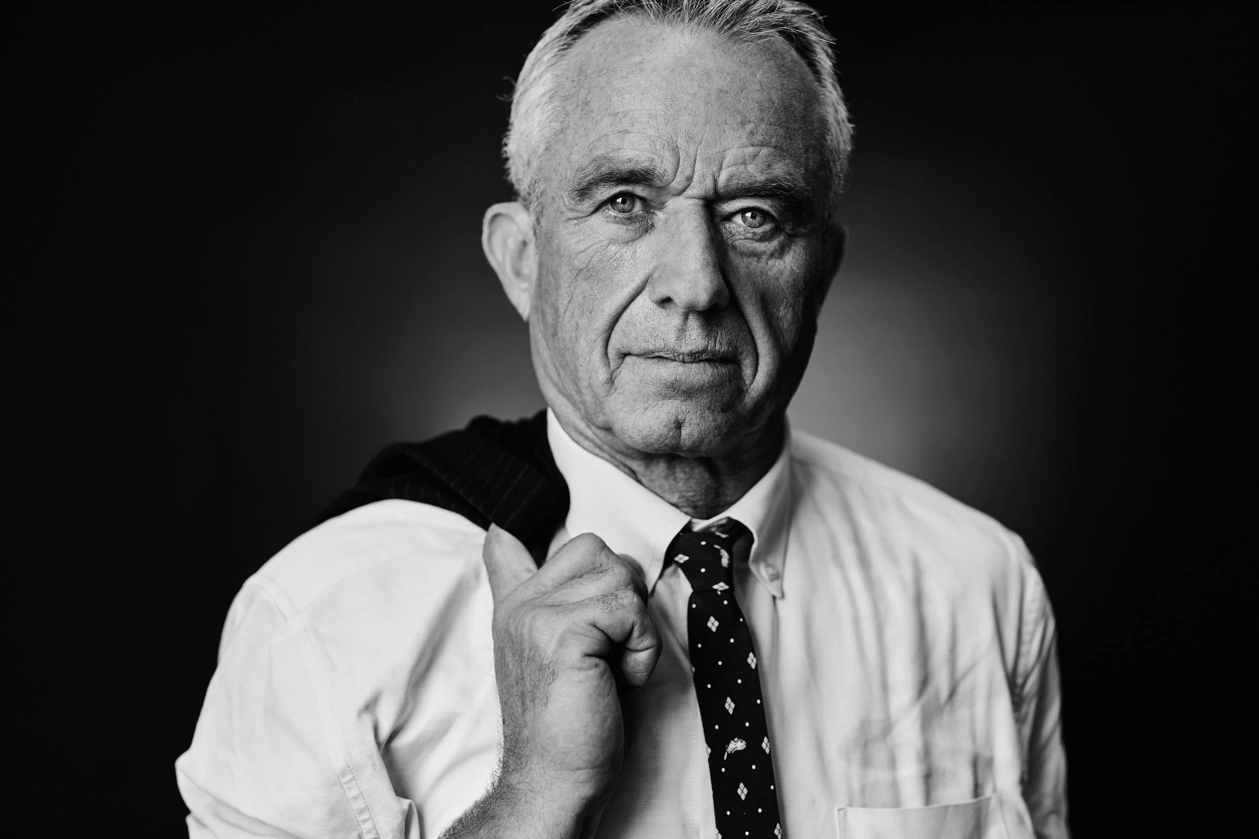 A black and white portrait of an elderly man with gray hair, wearing a white shirt and a dark tie with small patterns. He is adjusting his tie with his right hand, looking directly at the camera with a serious expression.