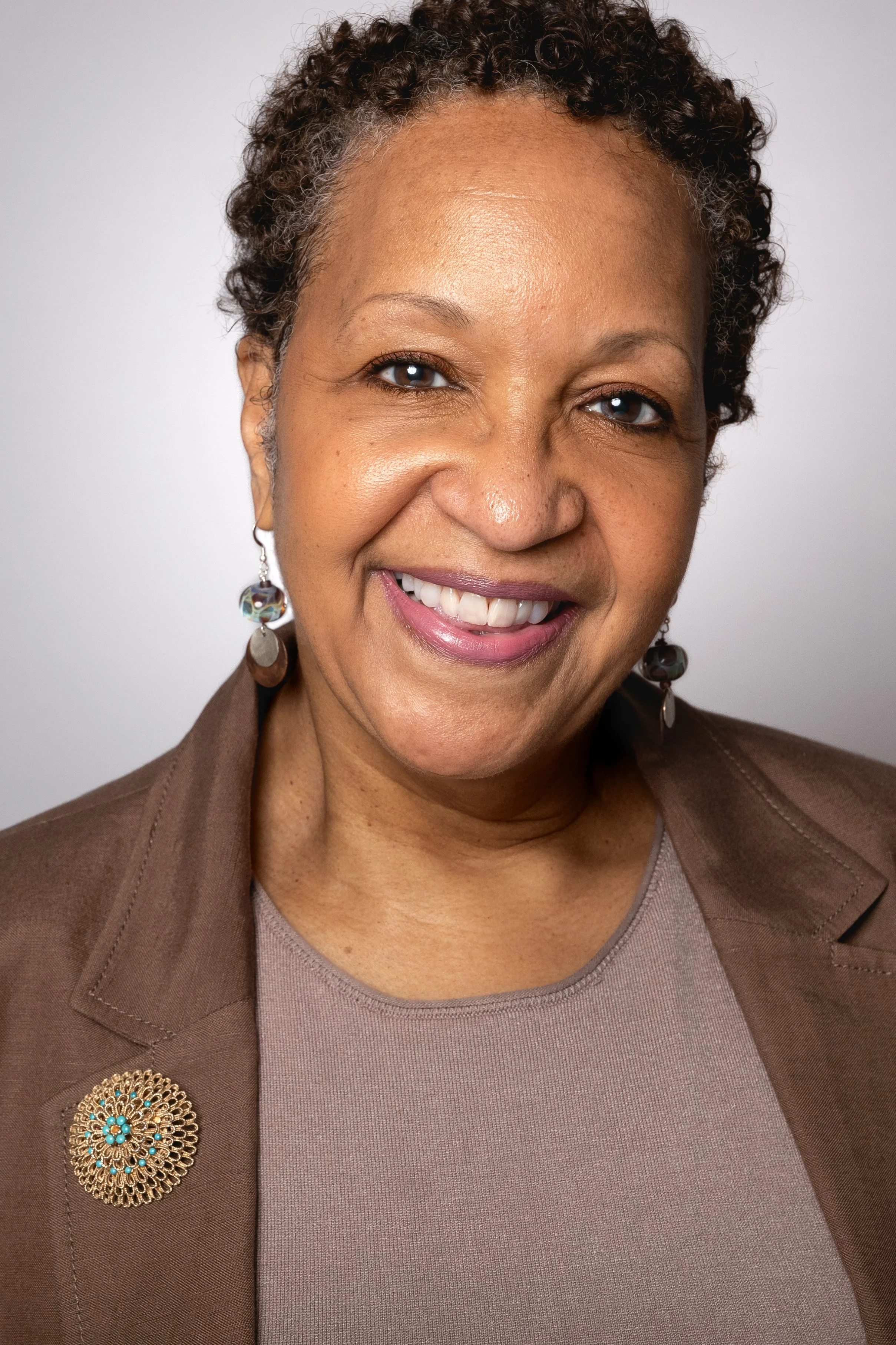 A smiling woman with short curly hair, wearing a brown blazer with a decorative brooch, earrings, and a taupe top against a plain white background.