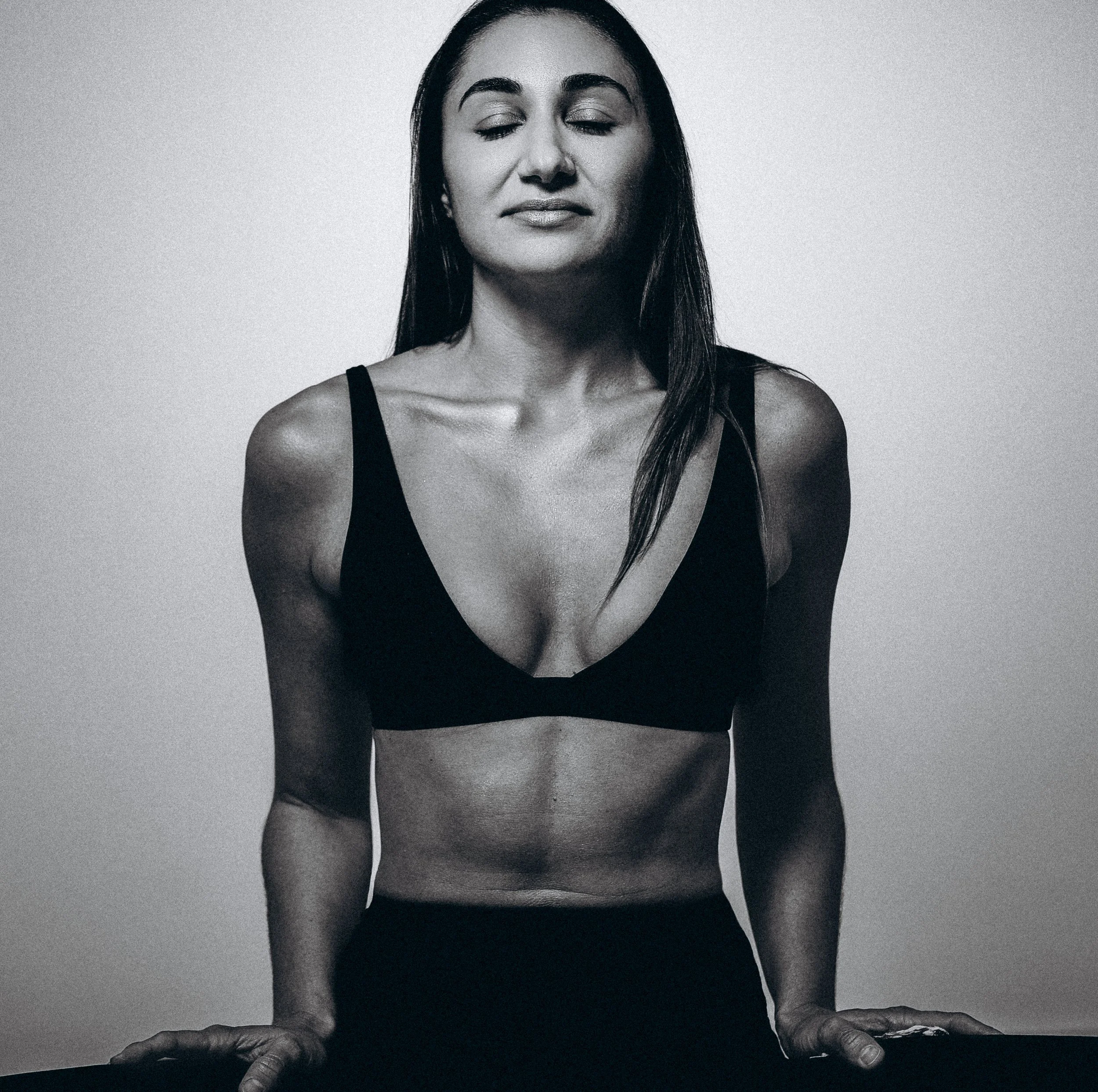 Black and white photo of a woman with closed eyes, long dark hair, wearing a black tank top, with her hands placed on a surface in front of her.