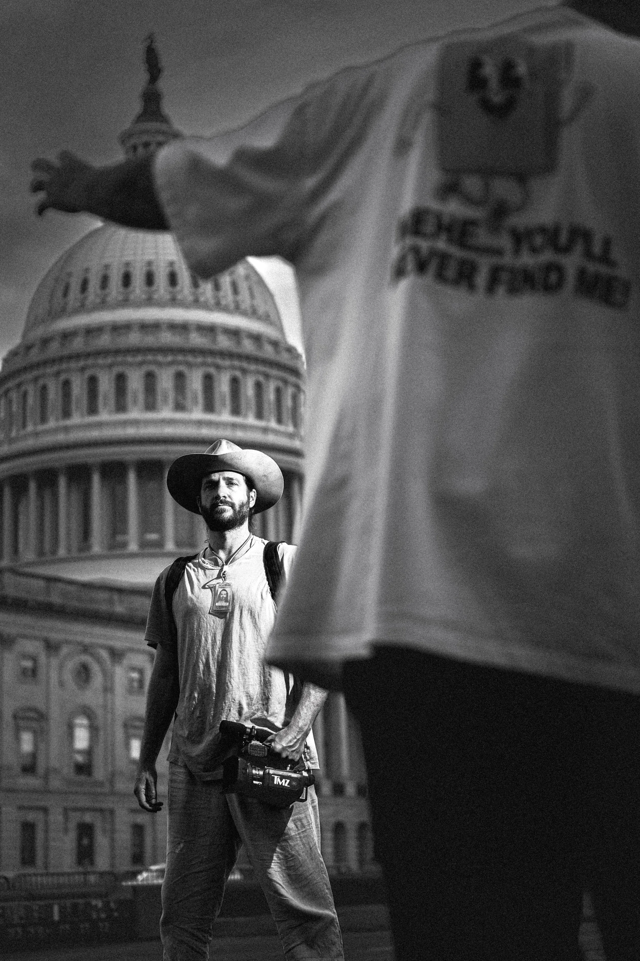 A man with a beard wearing a hat, a t-shirt, and a camera bag, standing in front of the U.S. Capitol building, with a partially visible person in the foreground and a flag waving.