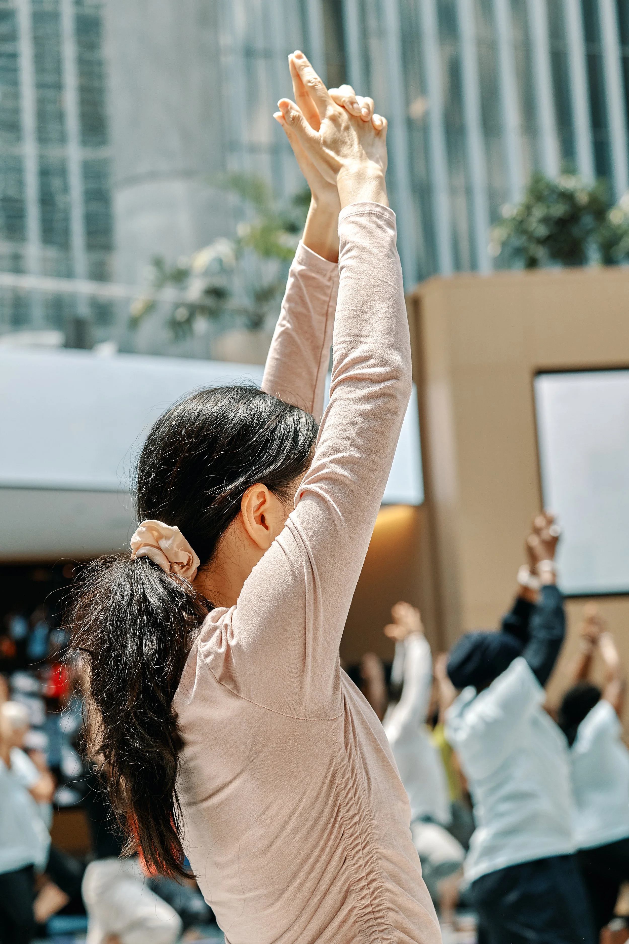 A woman with dark hair in a ponytail, wearing a beige long-sleeve top, raising her hands above her head during an outdoor event with a crowd, modern glass buildings and trees in the background.