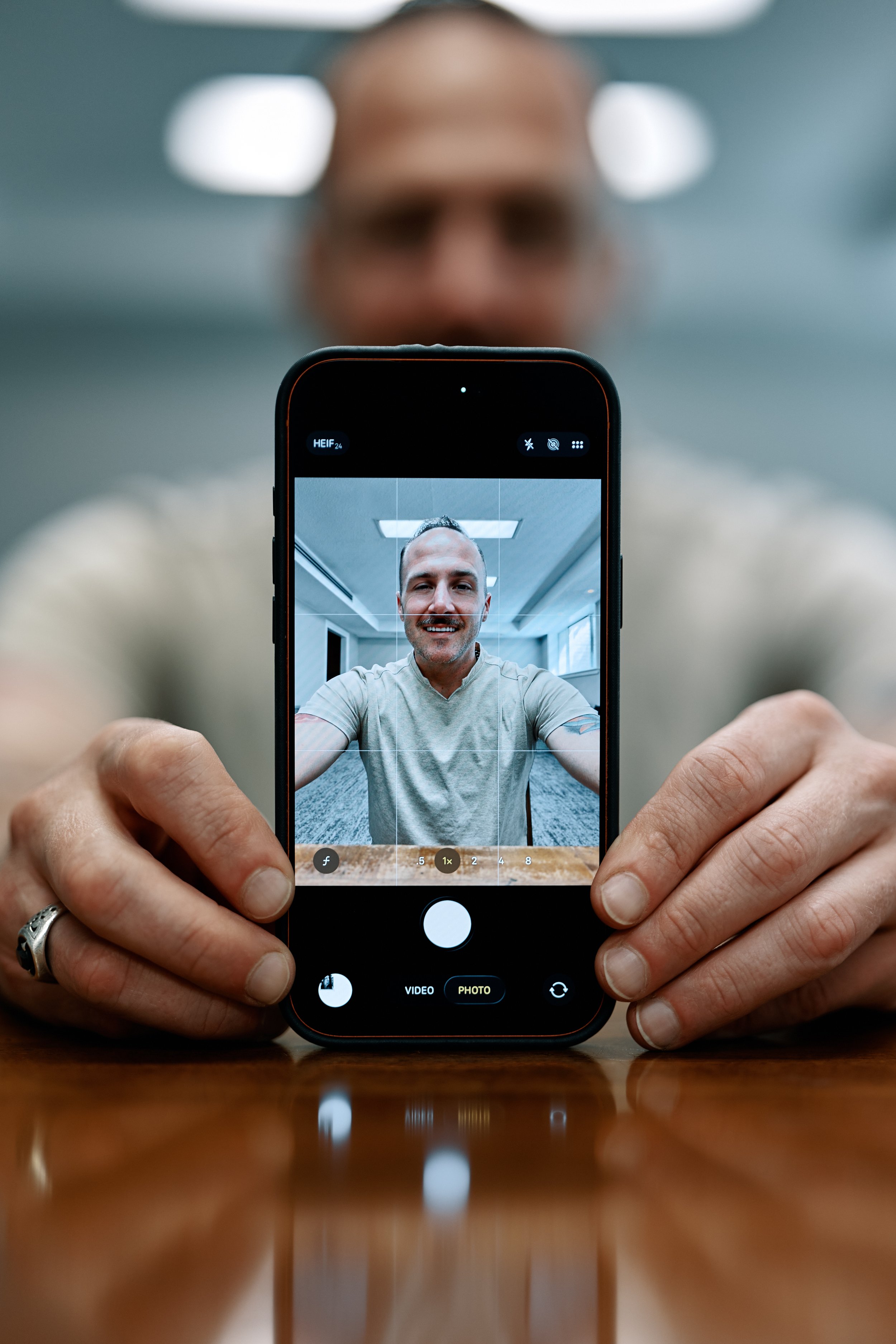 A person, Front Row Frankie, is taking a photo with a smartphone, capturing a smiling man sitting at a table in a well-lit room.
