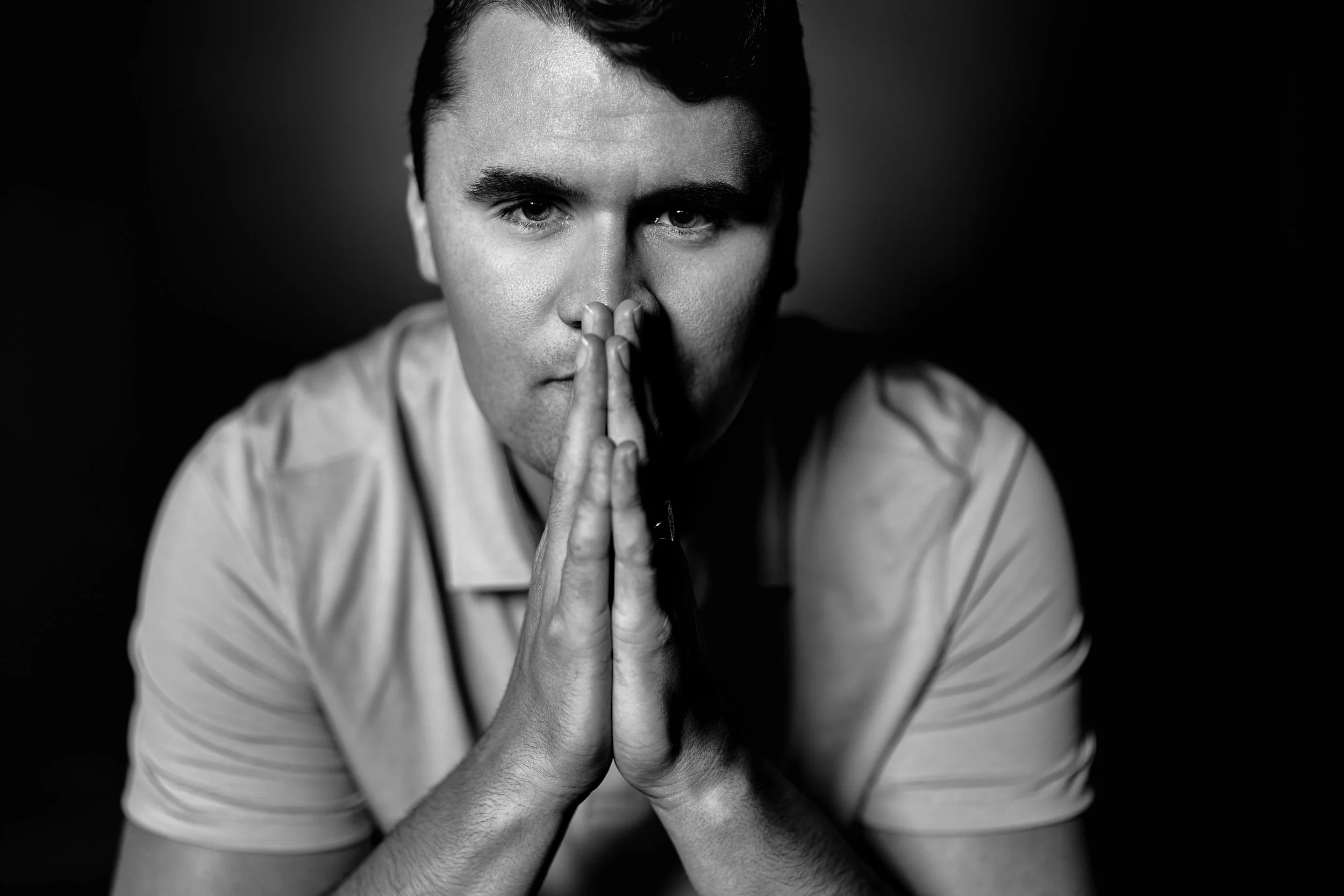 Black-and-white portrait of a man with short dark hair, pressing his hands together in front of his face, with a serious expression.
