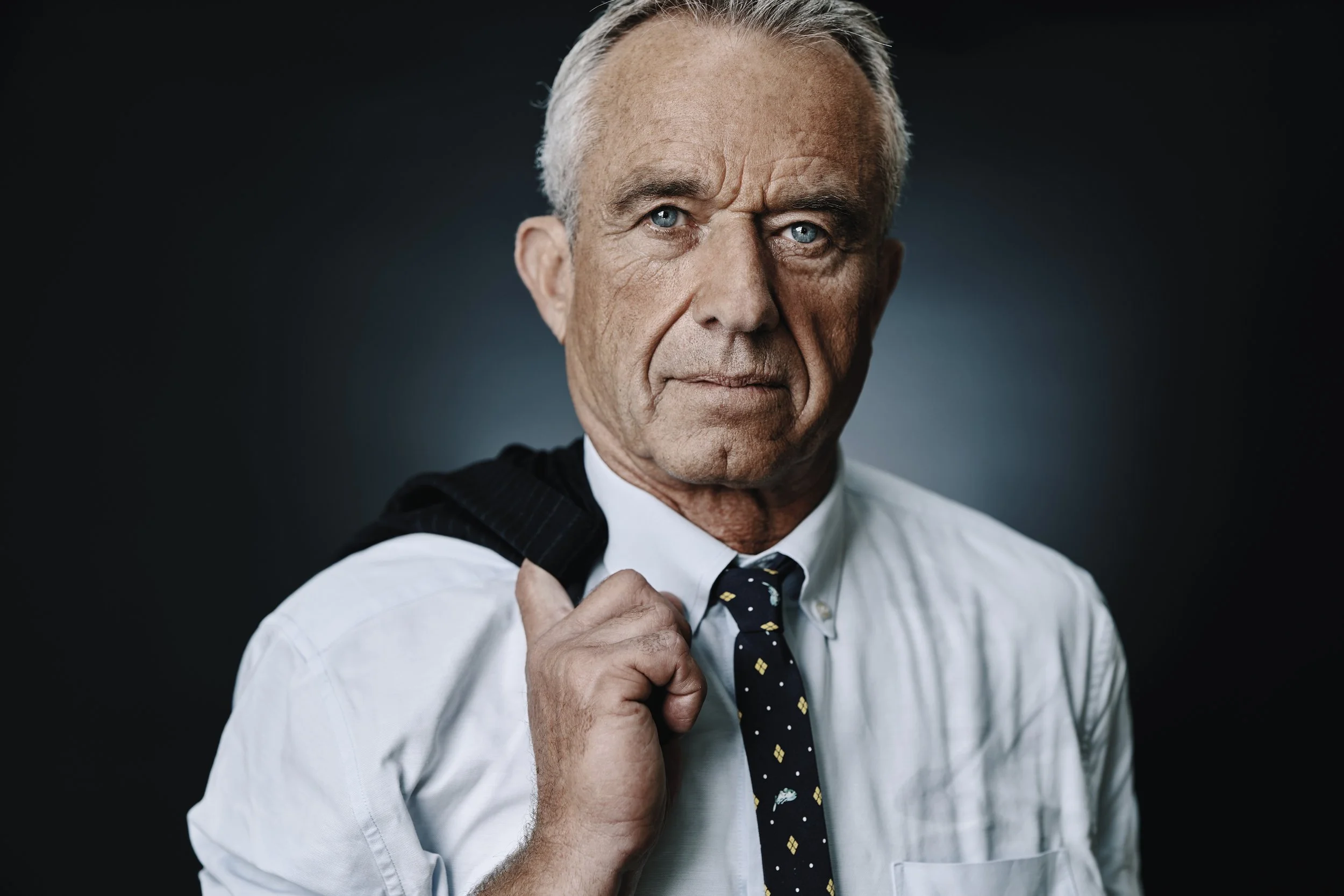 An older man with gray hair and blue eyes, dressed in a white shirt, holding a dark jacket over his shoulder and wearing a navy blue tie with yellow and white patterns, looking directly at the camera against a dark background.