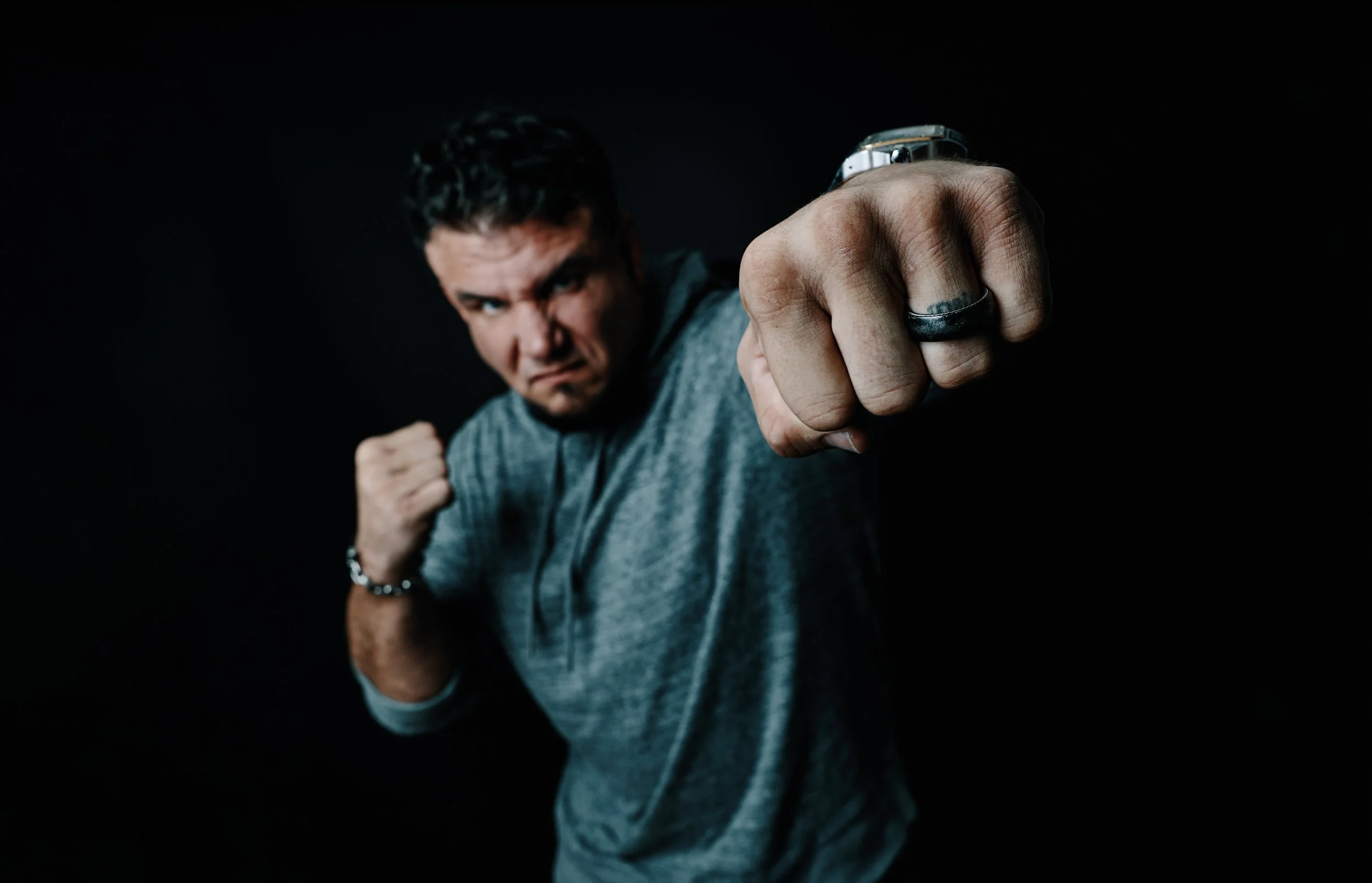 A man with dark hair and light skin is punching towards the camera, wearing a gray t-shirt and a silver watch. The background is black.