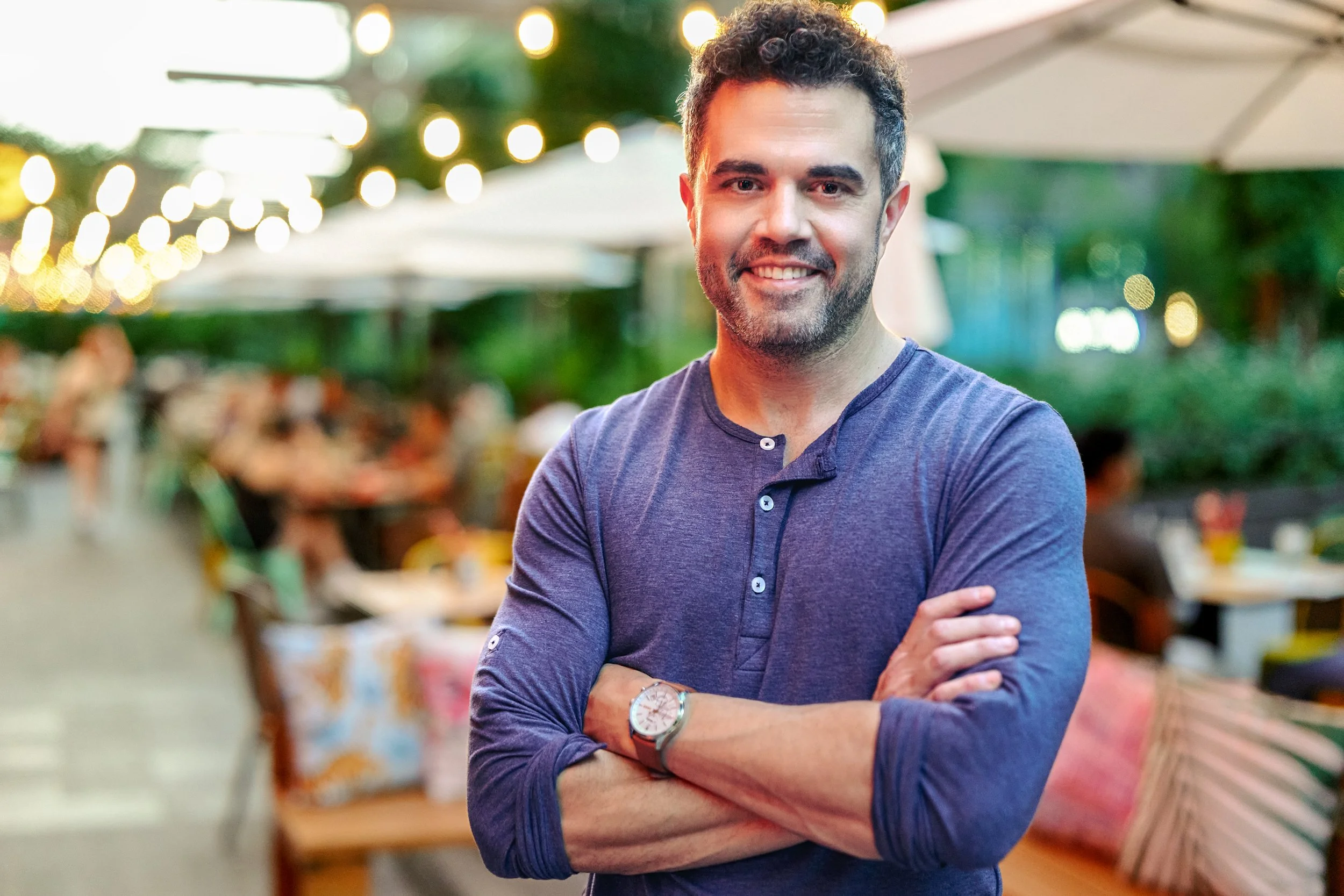 A man with dark curly hair, wearing a dark bluebutton-up shirt with rolled sleeves, standing with his arms crossed and smiling at camera in an outdoor setting with string lights and green trees in the background.