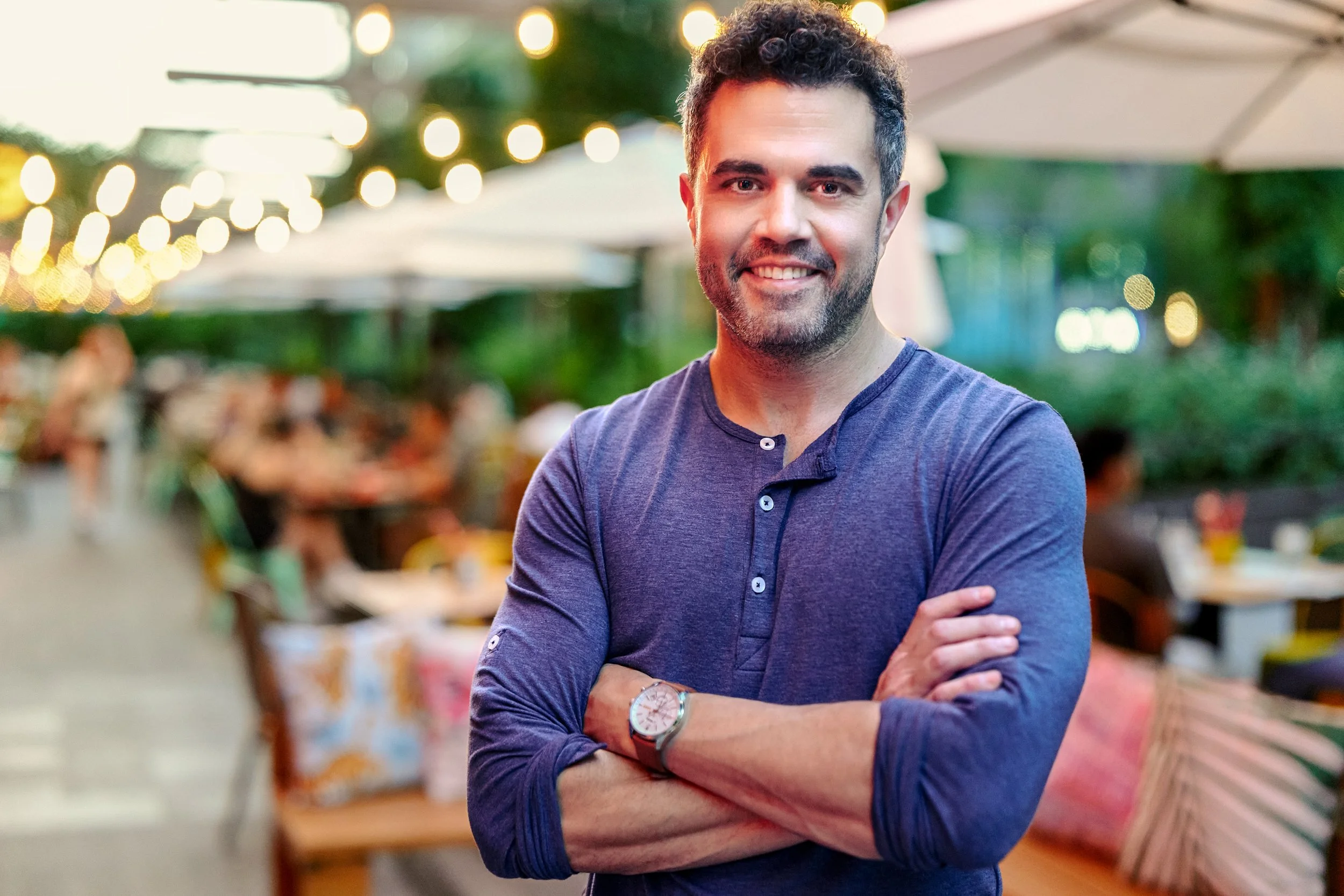 Young man with curly hair and a beard smiling at the camera, wearing a navy blue shirt with rolled sleeves in an outdoor setting with blurred lights and people in the background.