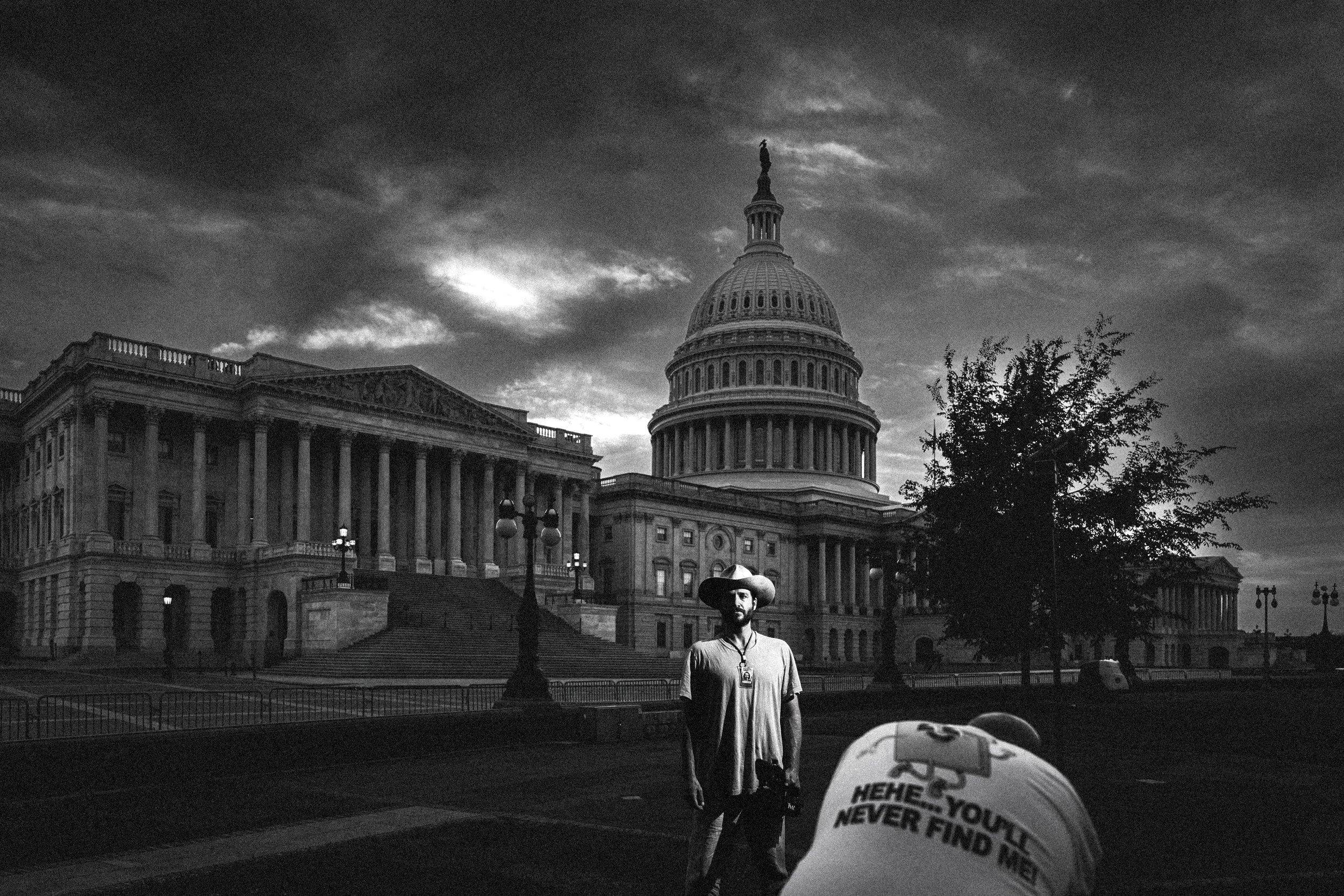 Black and white photo of the United States Capitol building, with a man wearing a wide-brimmed hat in the foreground. The sky is cloudy and there are some trees and street lamps visible.