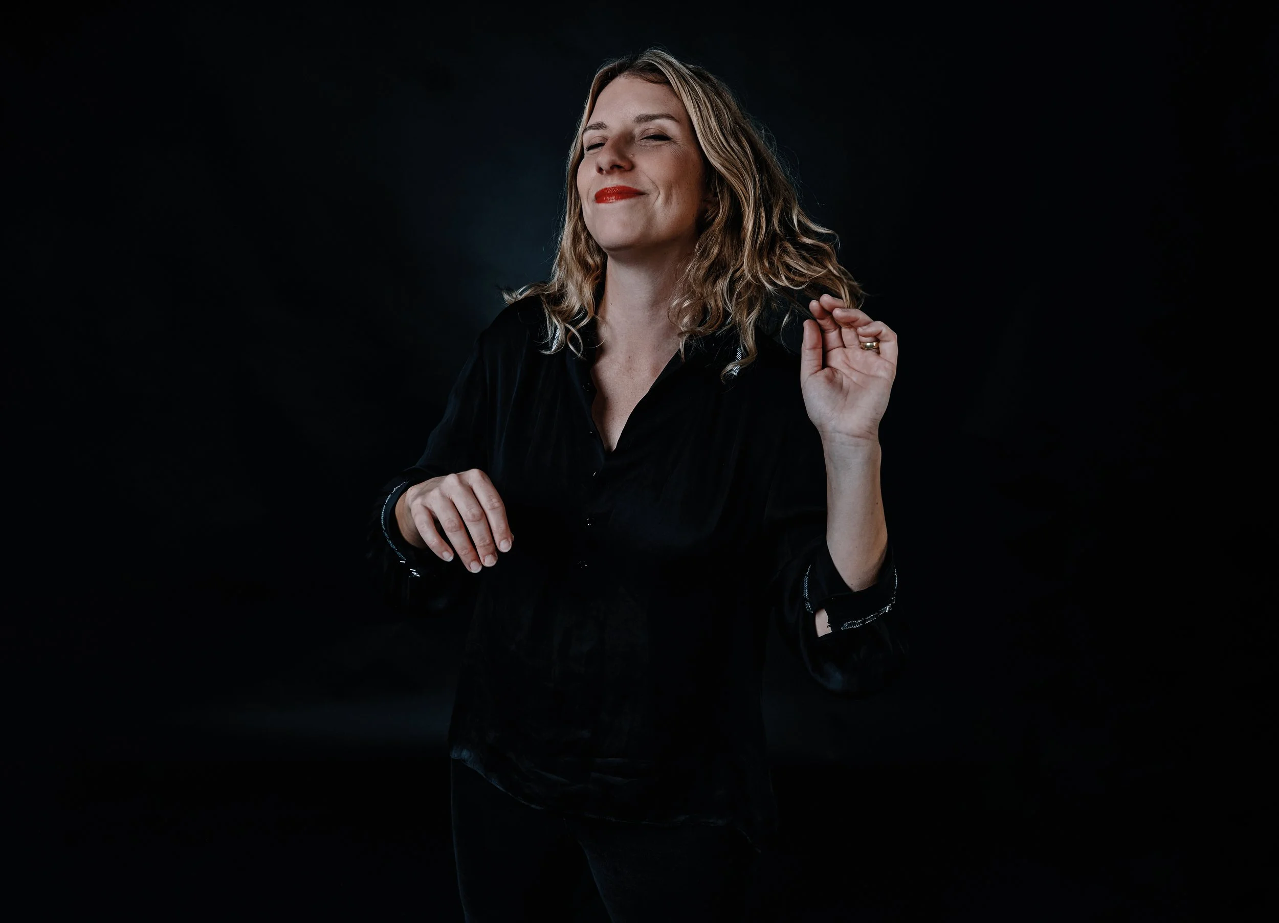 A woman, Leigh Brown, with wavy blonde hair, wearing a black shirt, is smiling with her eyes closed and holding her right hand near her face, against a dark background.