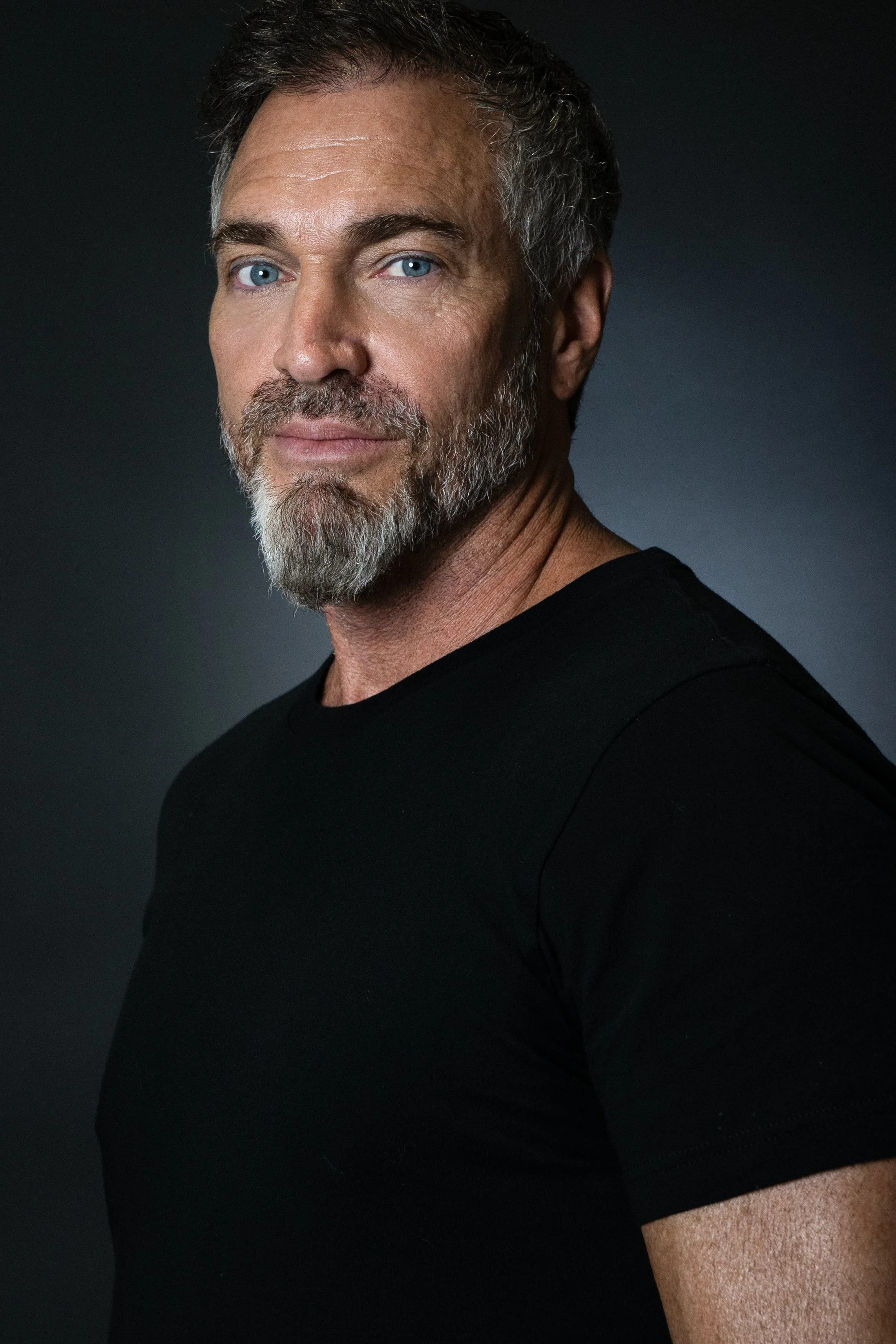 Close-up portrait of a middle-aged man with blue eyes, salt-and-pepper hair, and a beard, wearing a black shirt against a dark background.