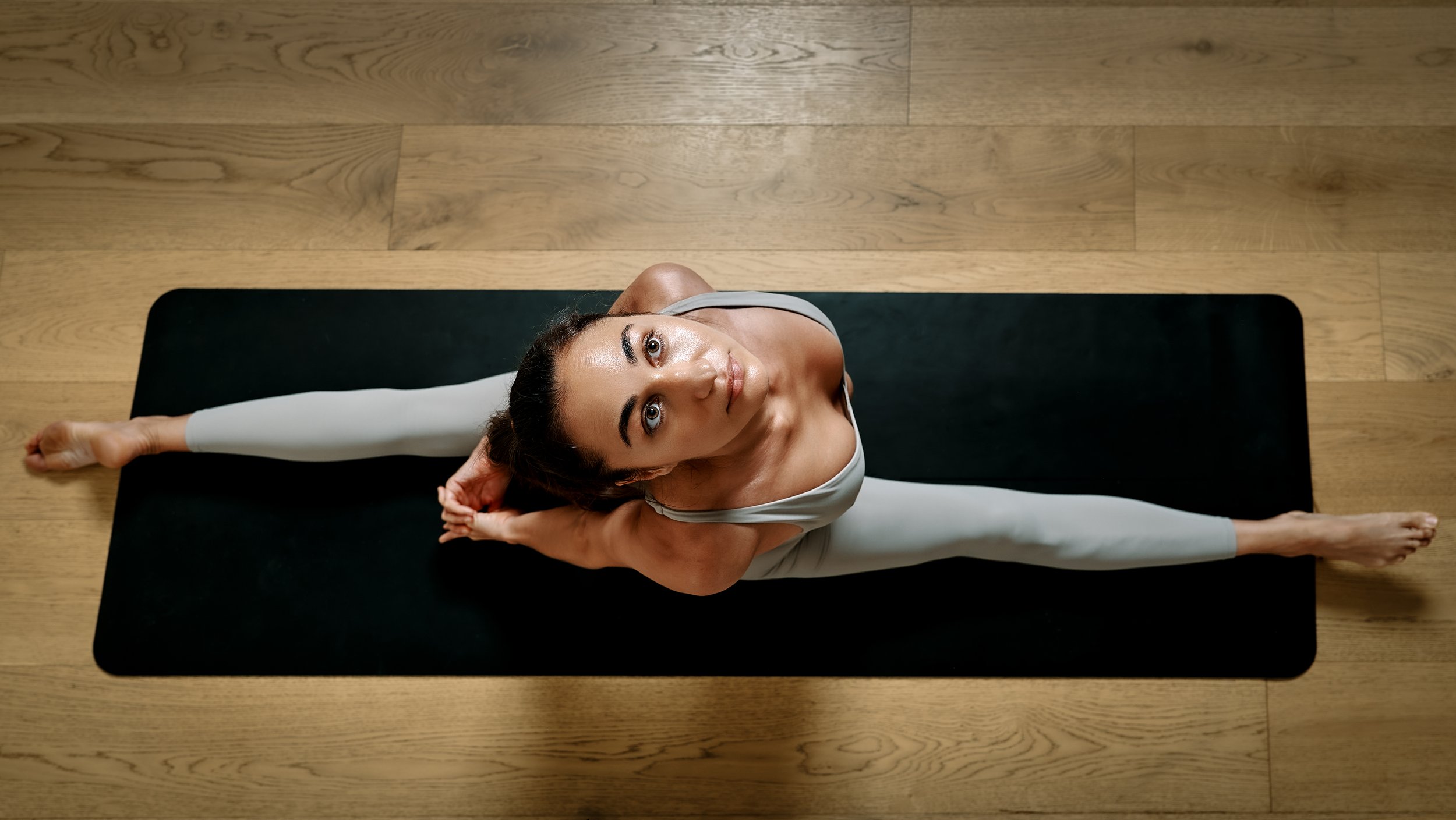 Woman practicing yoga indoors on a black mat on wooden flooring, doing a split stretch while looking up.