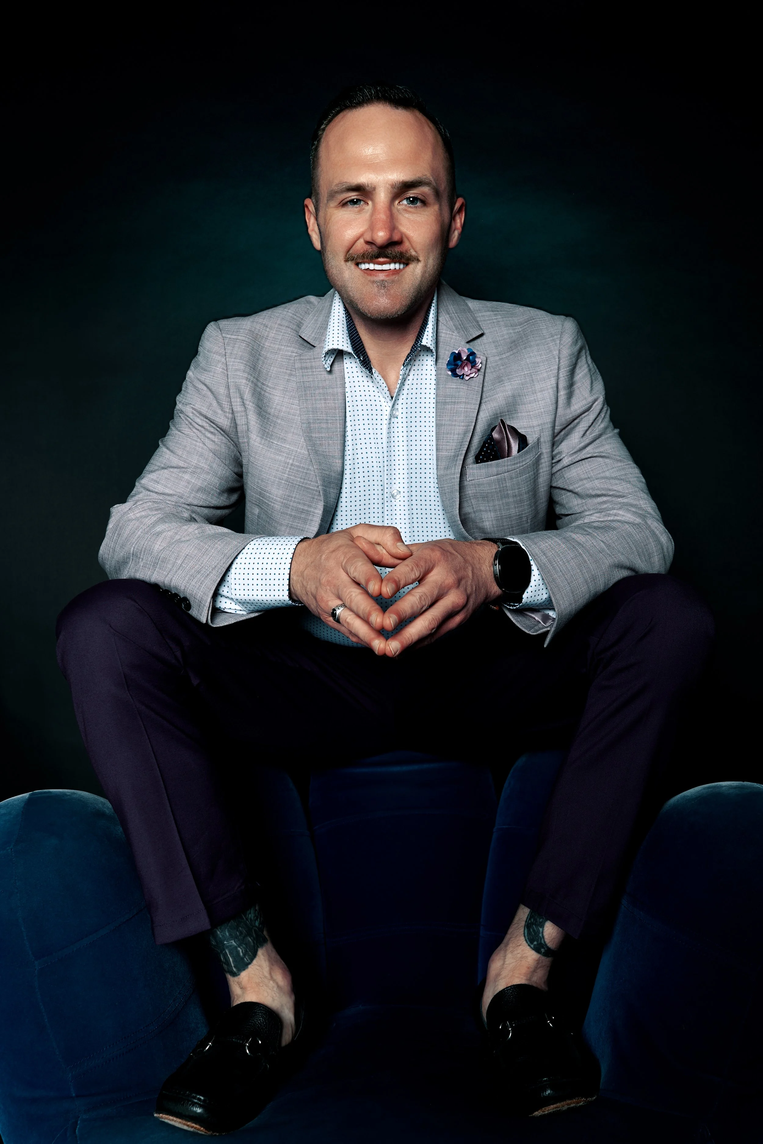 Man in a light gray blazer and white dress shirt with a small check pattern, sitting with his hands clasped, smiling, in front of a dark background.