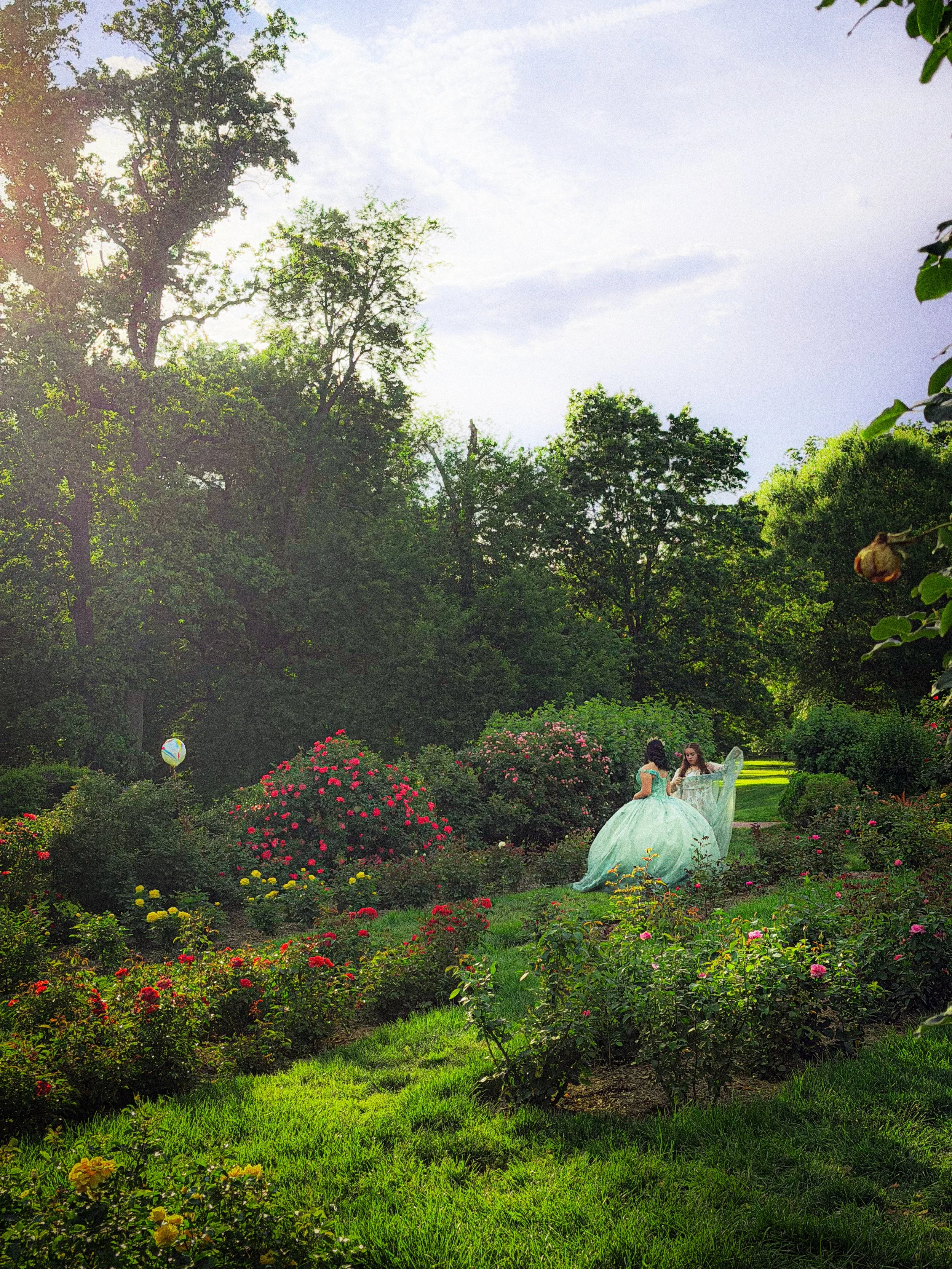 A garden scene with flowers, trees, and two women in vintage dresses, one with a large skirt, walking among the plants.