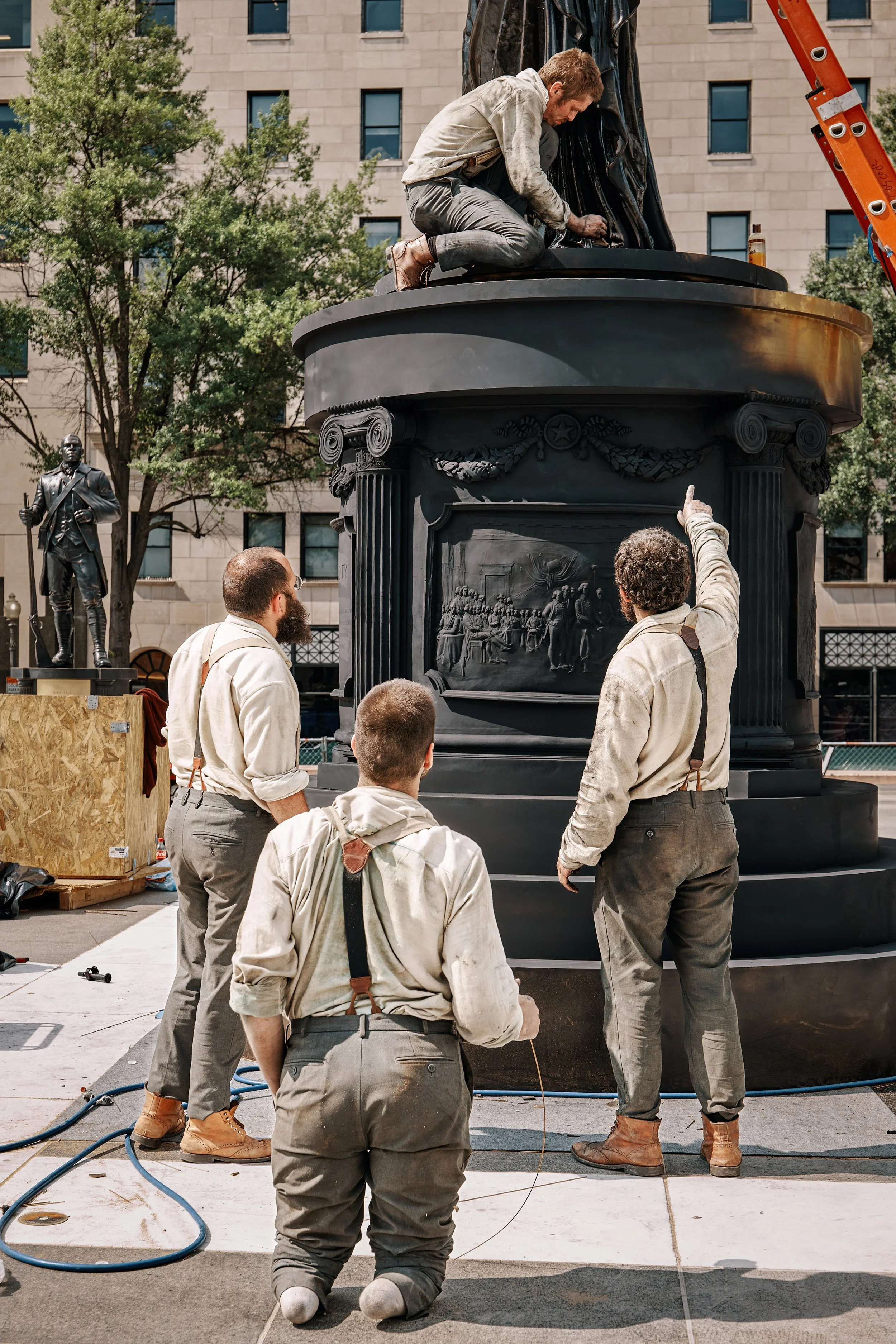French sculptors installing a large, black bronze statue on a platform outdoors, with trees and a beige building in the background at Freedom Plaza in Washington, DC. 