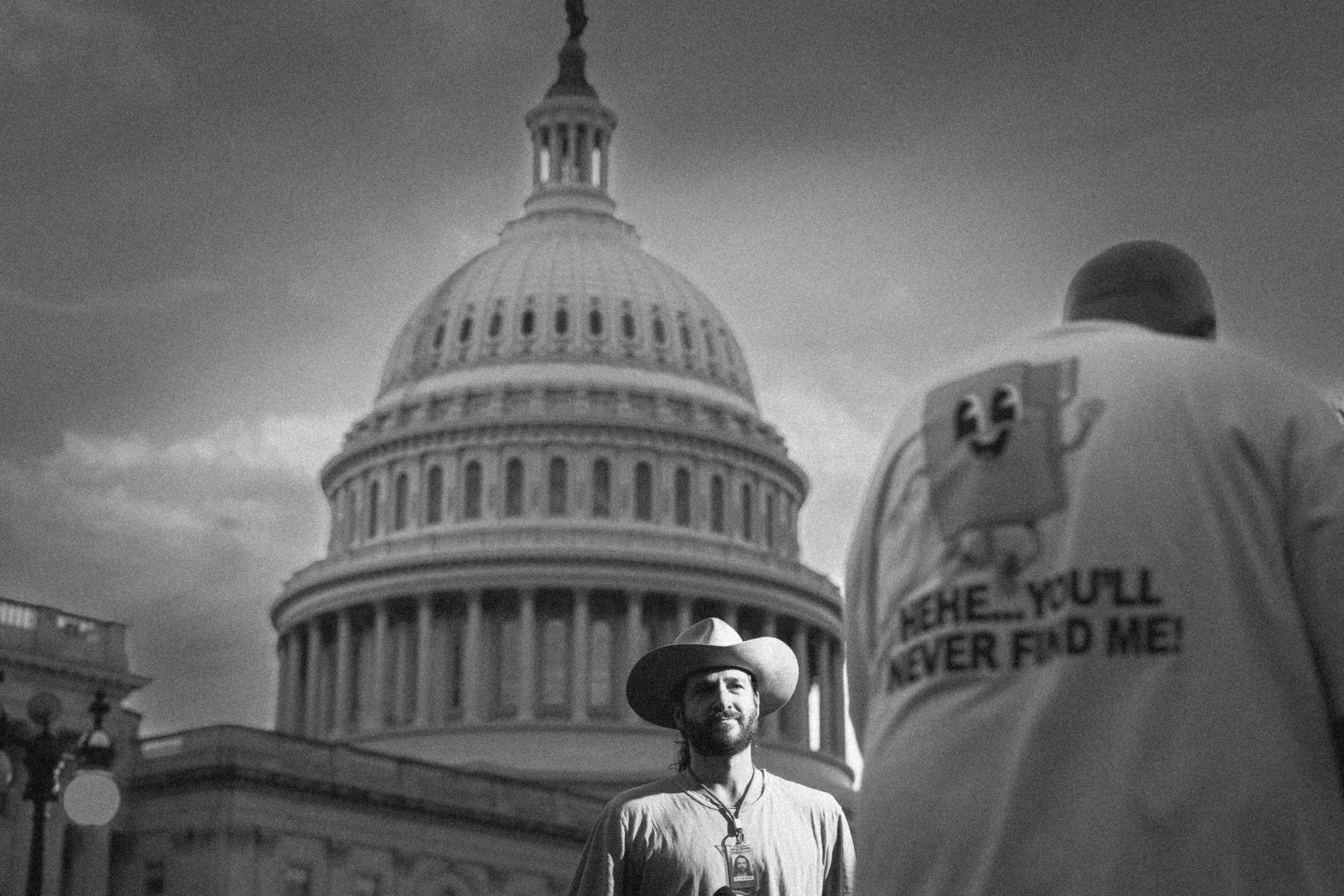 A man wearing a wide-brimmed hat and a badge around his neck stands in front of the U.S. Capitol building. The photo is in black and white.