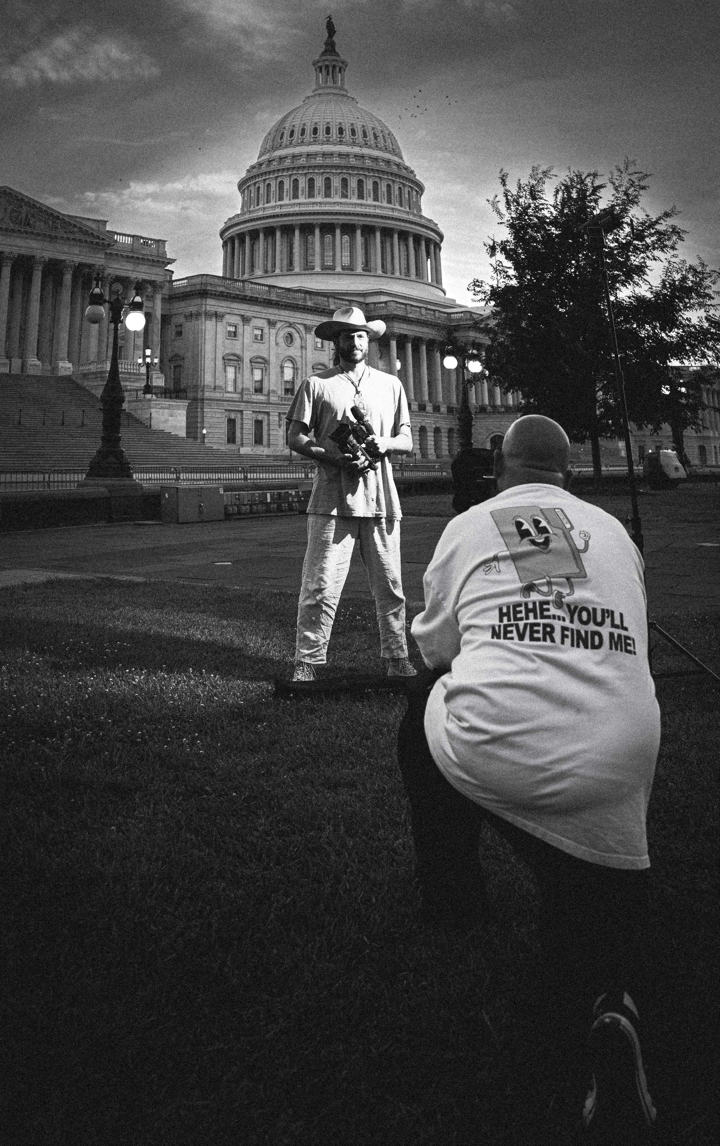 A black and white photo of two men in front of the U.S. Capitol building. One man is standing, holding a camera, and wearing a hat and casual clothes. The other man, kneeling nearby, wears a T-shirt with a cartoon character and the text, "Hehe... You