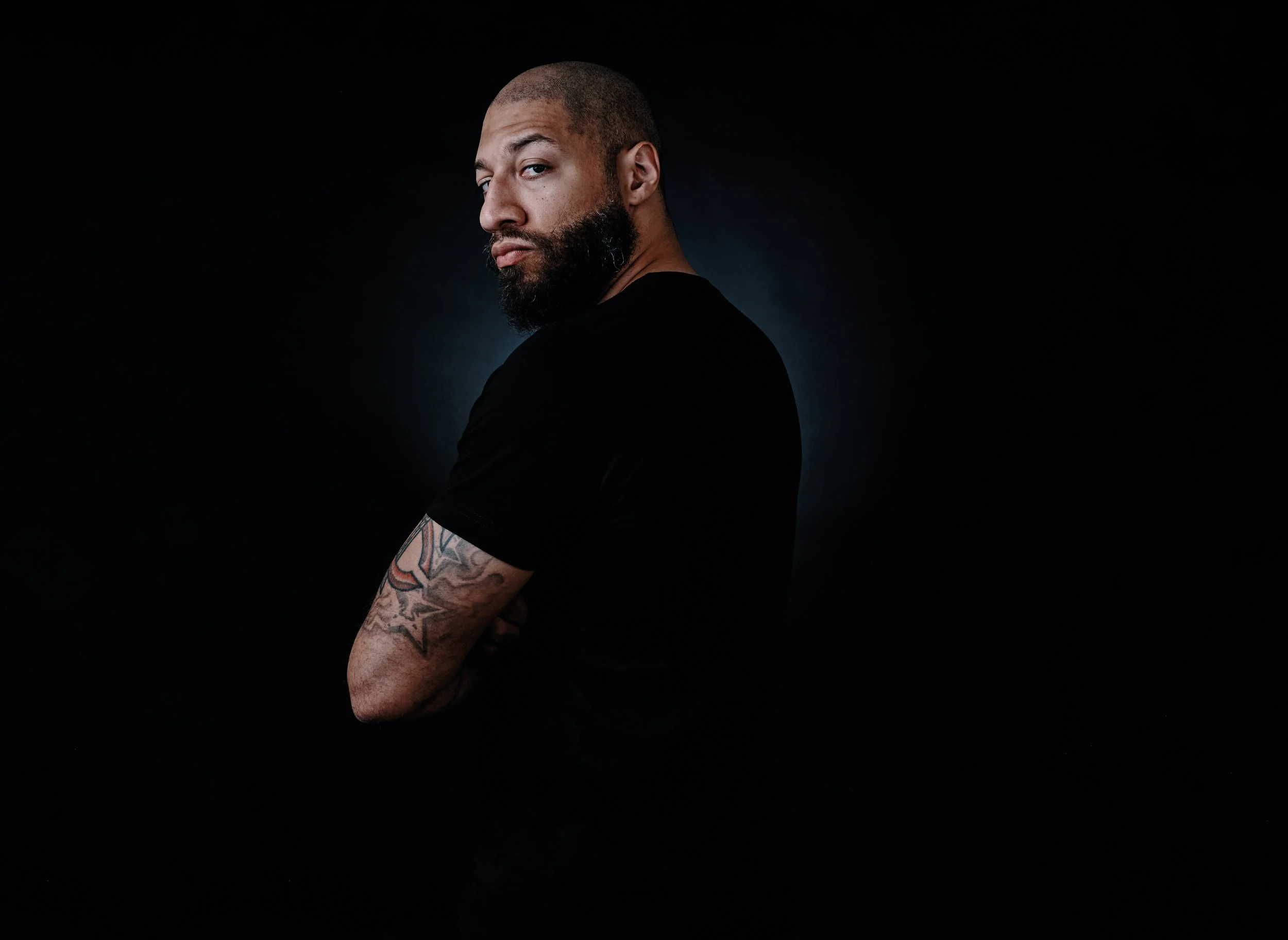 A man, former NBA player Royce White, with a beard and tattoos on his arm wearing a black shirt, standing against a dark background, looking over his shoulder with a serious expression.