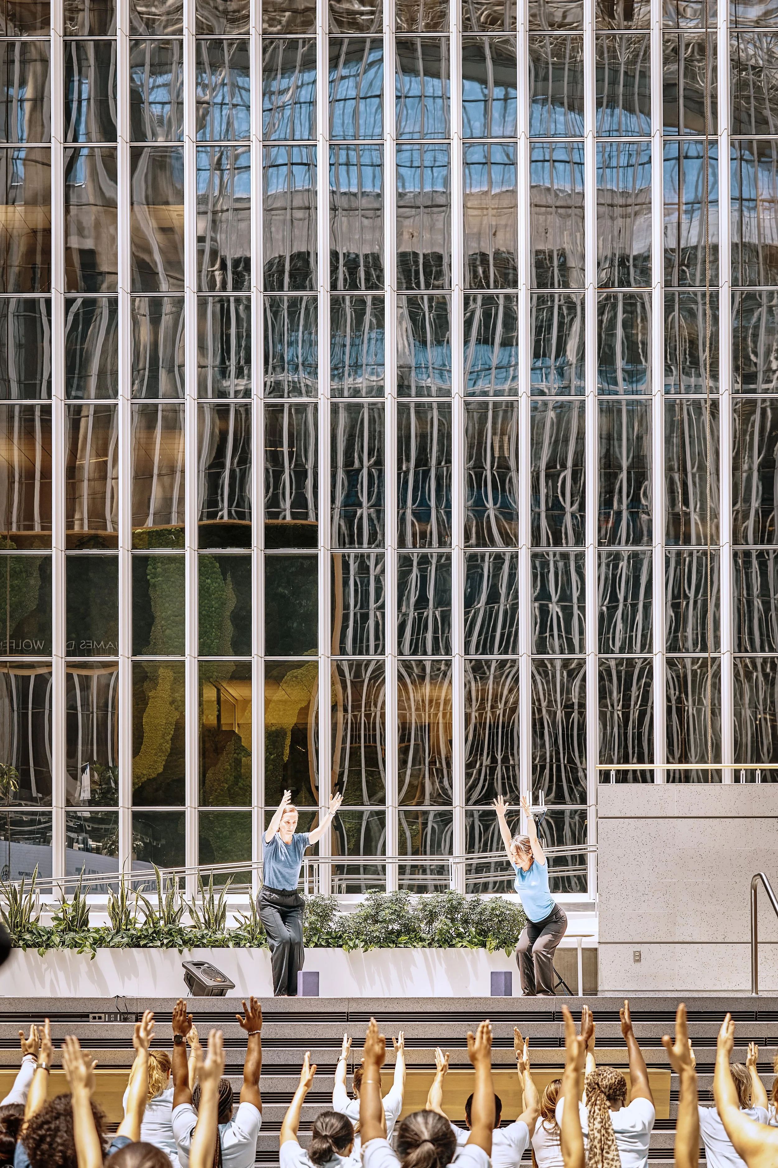 Two women perform yoga poses on an outdoor stage in front of a large, reflective glass building with distorted reflections. The audience in front of them has raised hands.