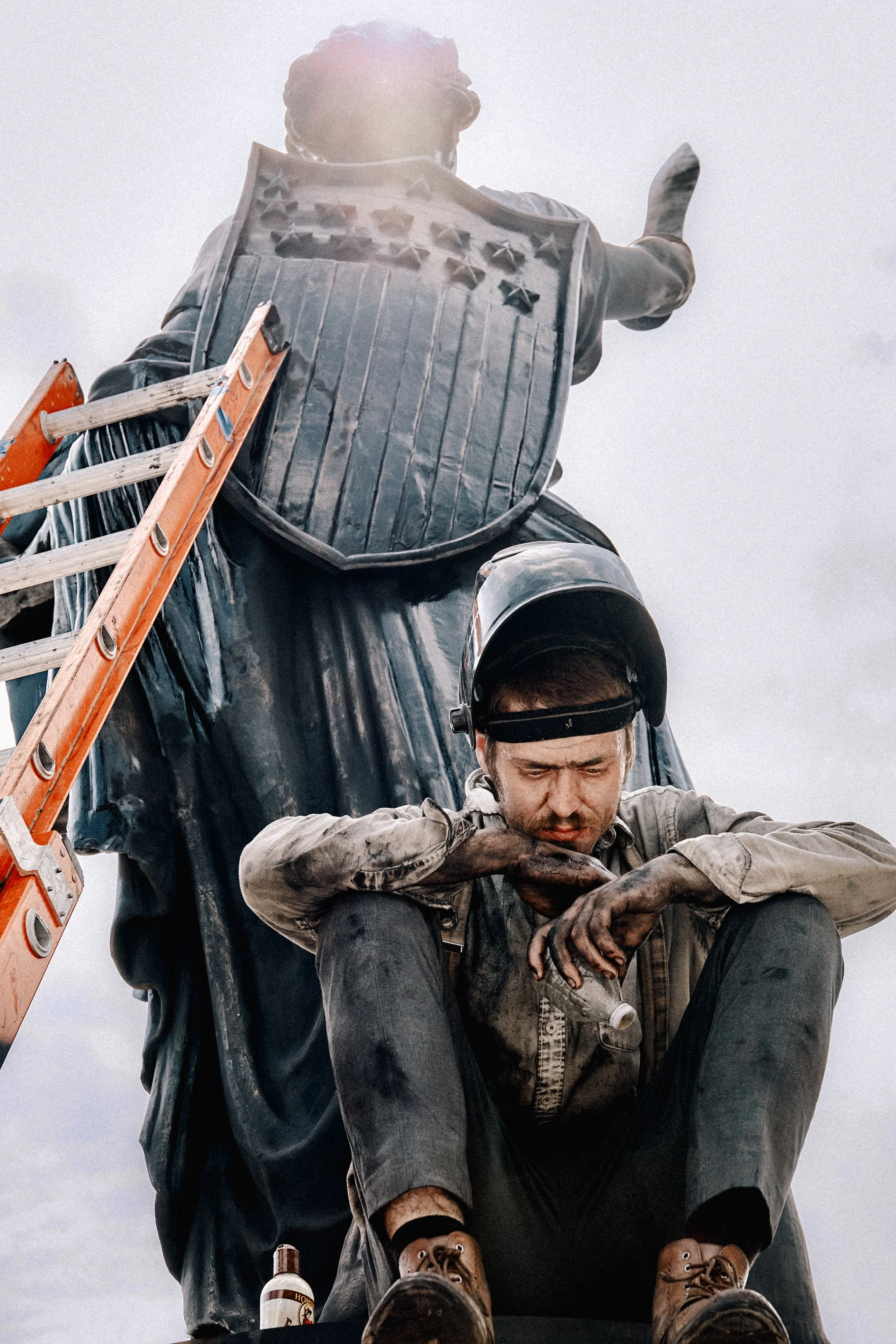 A man wearing a helmet sits on the ground with his head bowed, hands on his knees, and dirty clothing. Behind him, a statue of a soldier in uniform, with a shield and stars on it, is positioned as if watching over him. An orange ladder leans against 