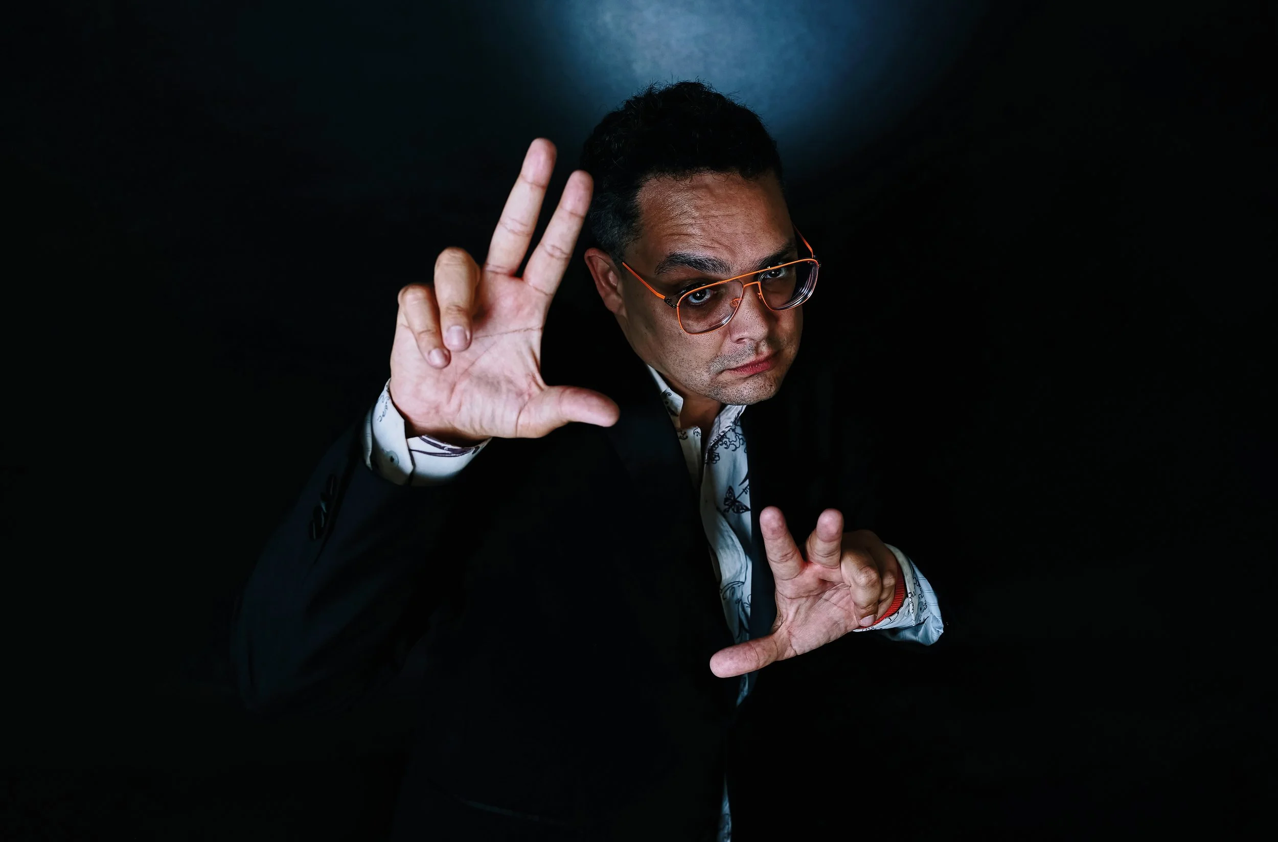 A man in glasses and a suit making peace and heart hand gestures against a dark background.