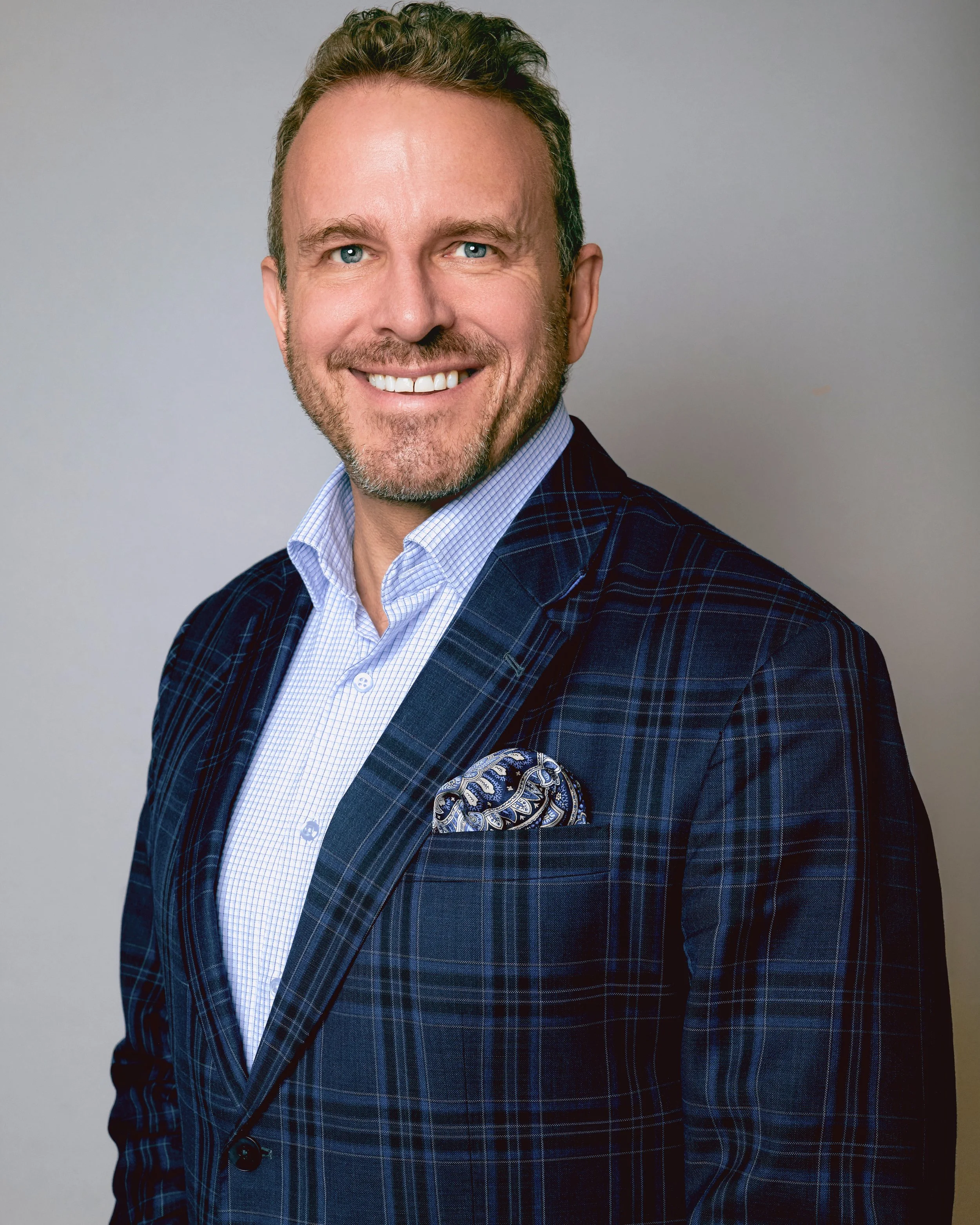 A man with light skin, blue eyes, and light brown hair is smiling and dressed in a blue checkered jacket, a light blue collared shirt, and has a patterned pocket square. The background is plain and gray.