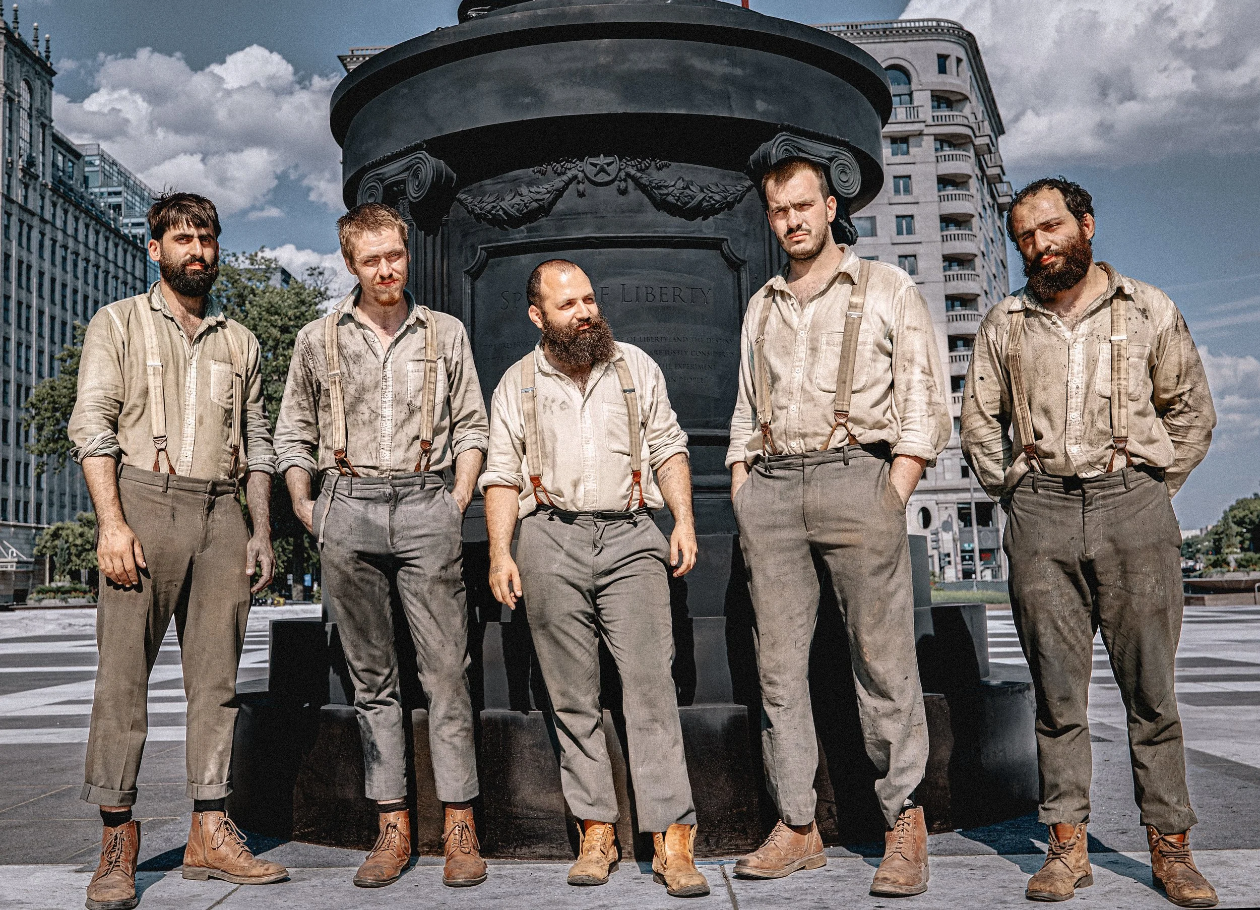 Five French sculptors in vintage work clothes with suspenders and boots standing in front of a large Statue of Liberty base in an urban area. Freedom Plaza, Washington, DC. 