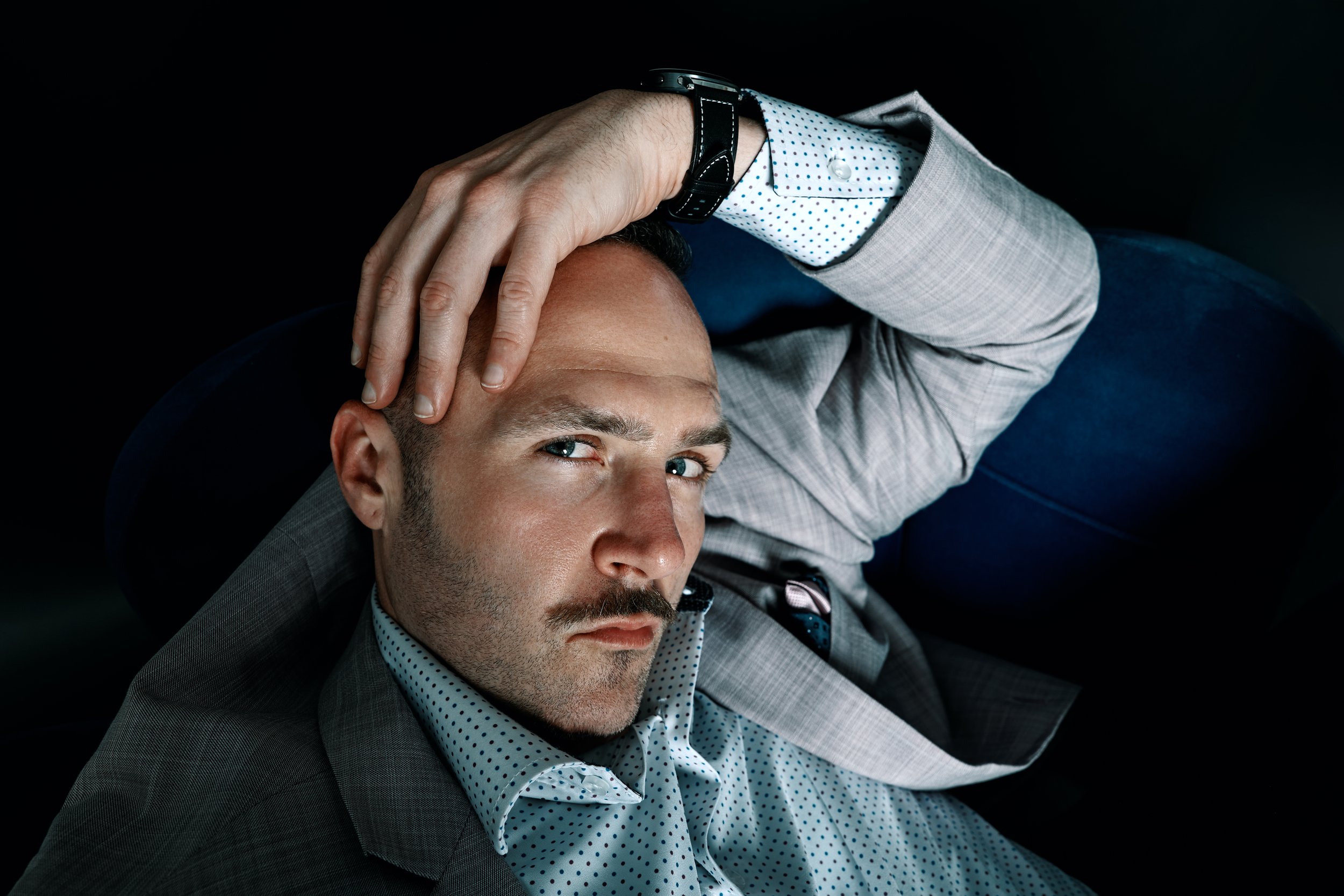 A man, Front Row Frankie, with light skin, short hair, and a beard,leaning back in a dark setting. He is wearing a gray plaid blazer, a white dress shirt and a black watch. His left hand is placed on his forehead as he looks directly at the camera.