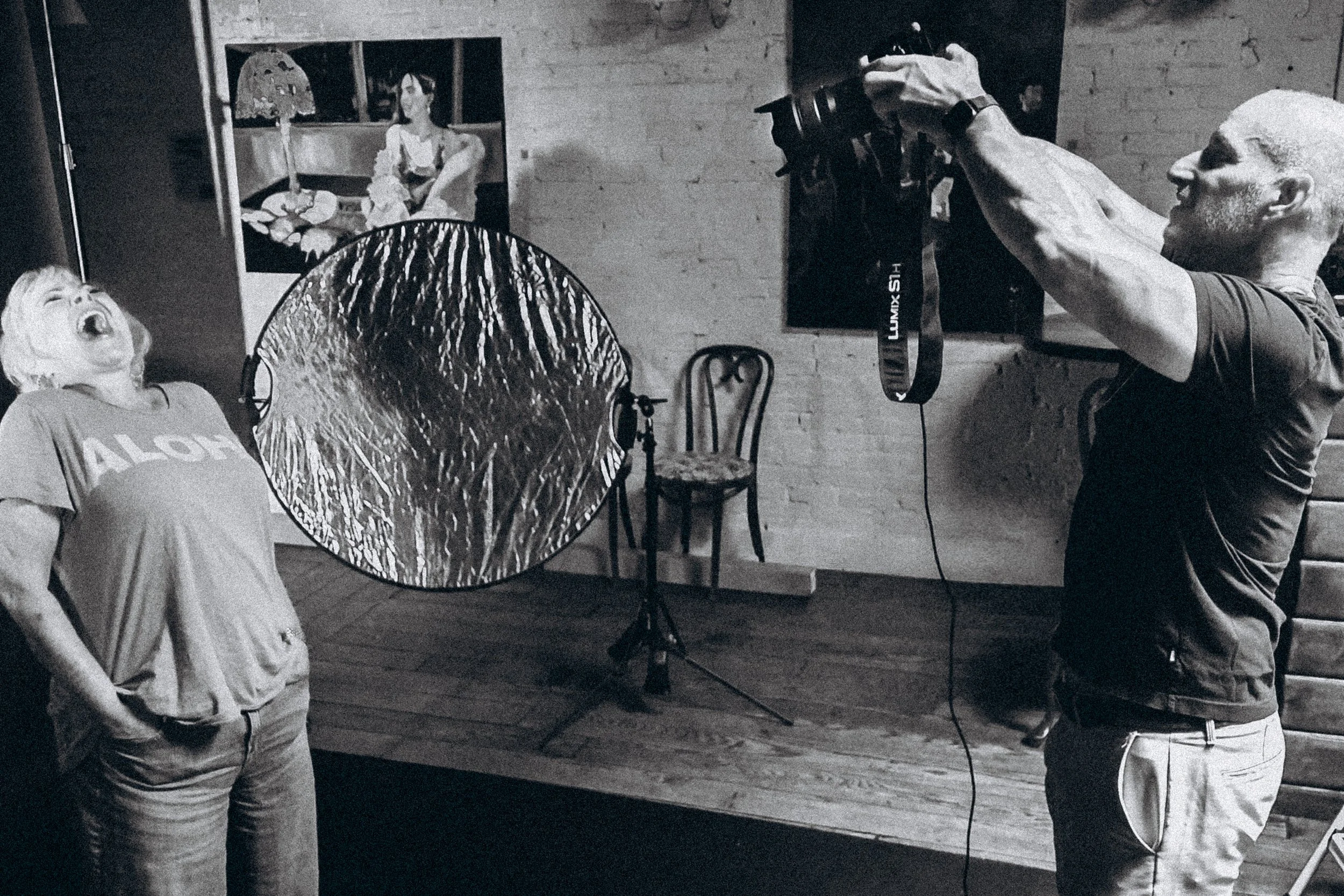 A man with a bald head and beard taking a photo of a woman laughing, both are indoors near a brick wall, with a large reflector and a framed painting of a woman sitting at a desk in the background.