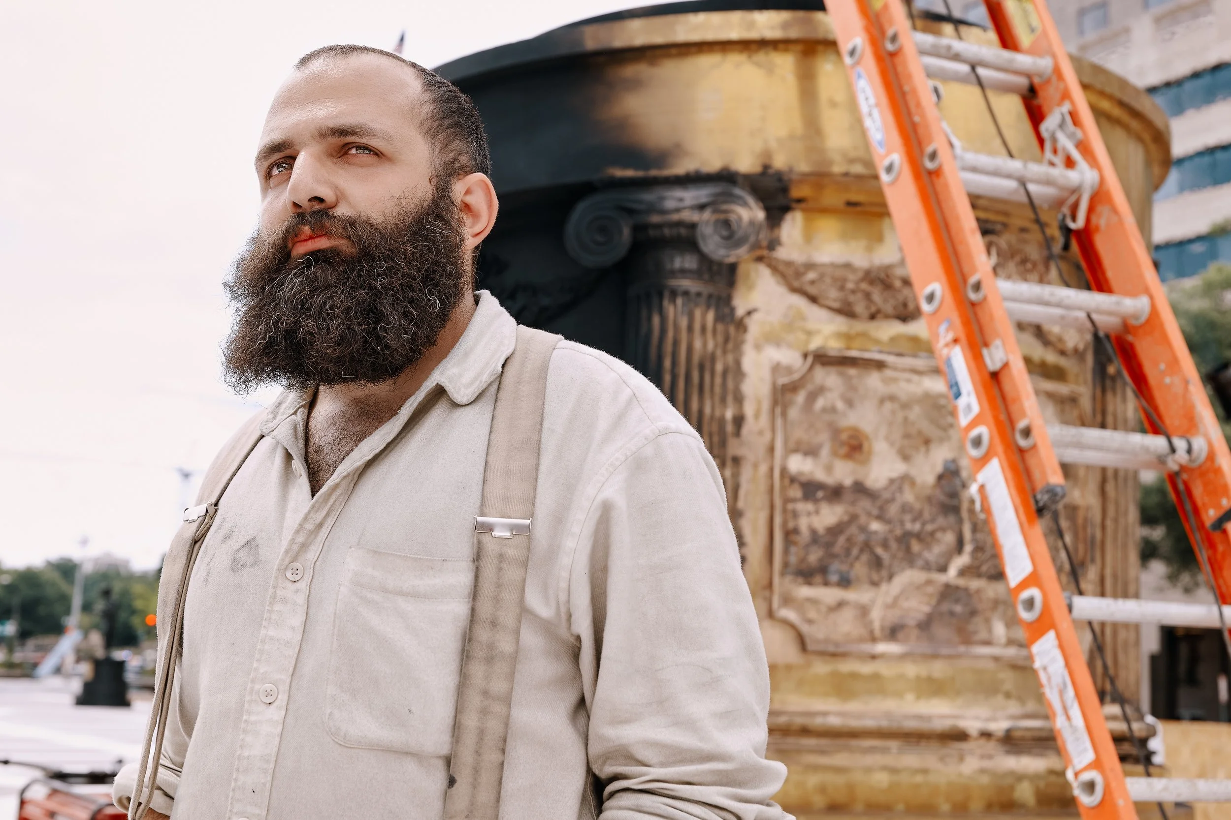 A French sculptor with a beard wearing a light-colored shirt stands outdoors at a sculpture installation site with a ladder and building materials behind him.