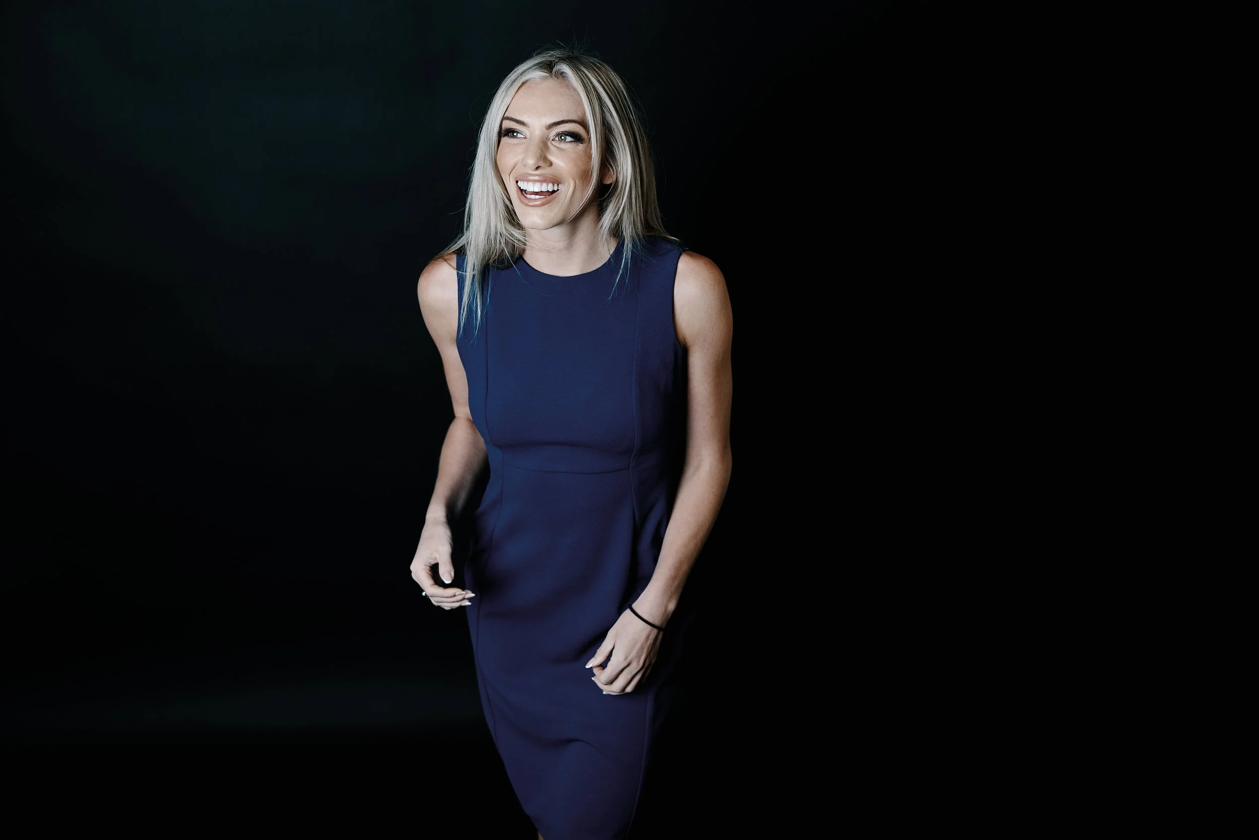 A woman with shoulder-length blonde hair wearing a sleeveless navy blue dress, standing against a black background, smiling, and looking to her left.