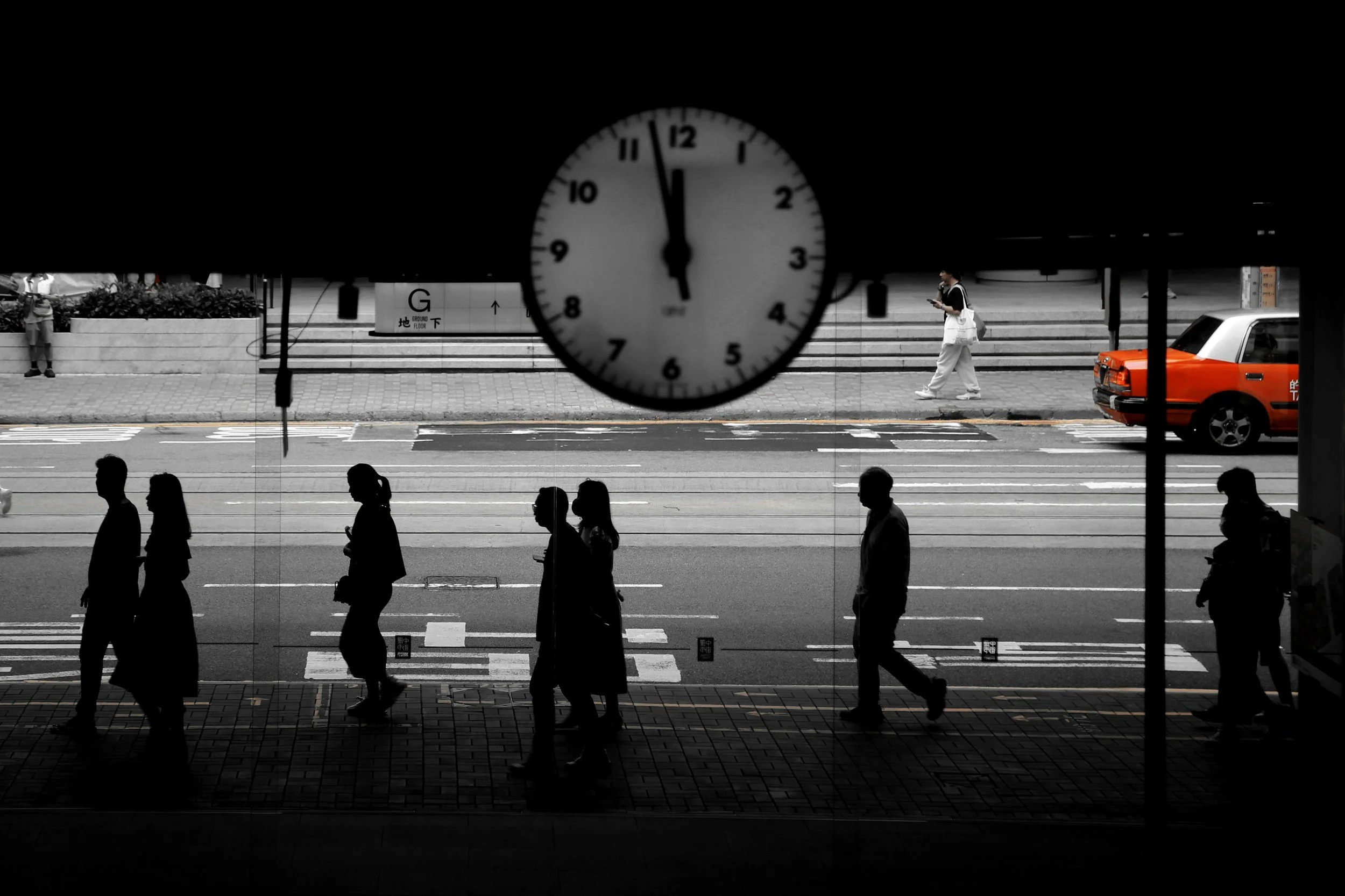A street scene viewed from inside a building, with silhouettes of pedestrians walking on the sidewalk. In the background, a clock shows nearly 12:55, and a person is seen crossing the street. Cars, including an orange taxi, are parked or driving by.