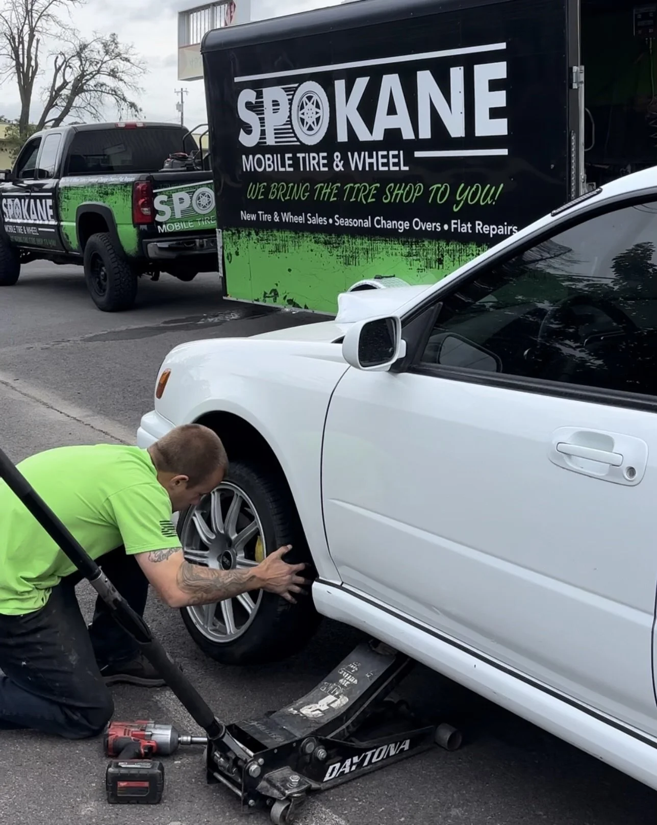 A person in a neon green shirt is changing or inspecting the wheel of a white car with a hydraulic lift underneath. In the background, a truck with a sign advertising a mobile tire and wheel service is parked on the street.