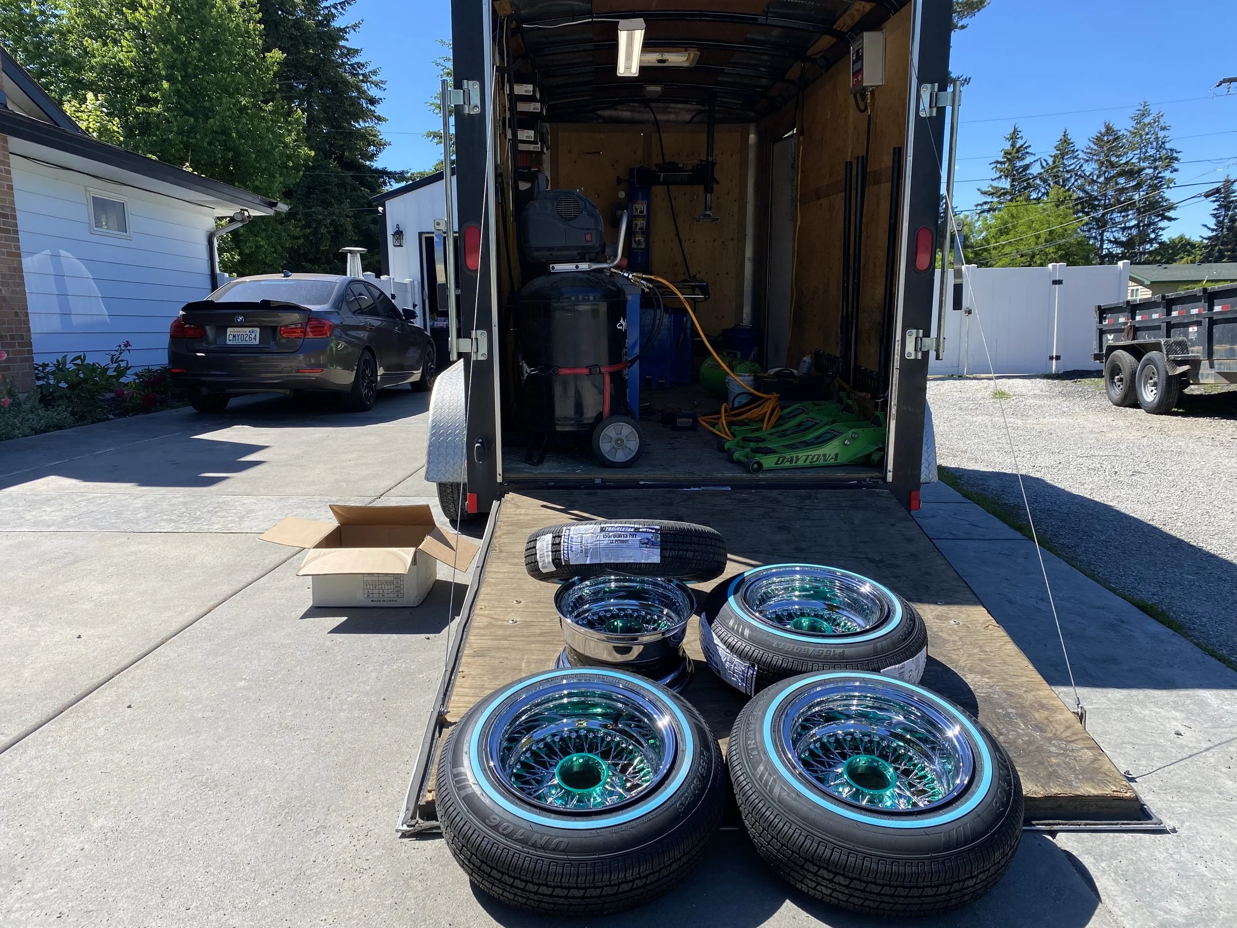 Open trailer with racing wheels, tire, and tools inside, parked on driveway next to a black car and a residential house with trees in the background.