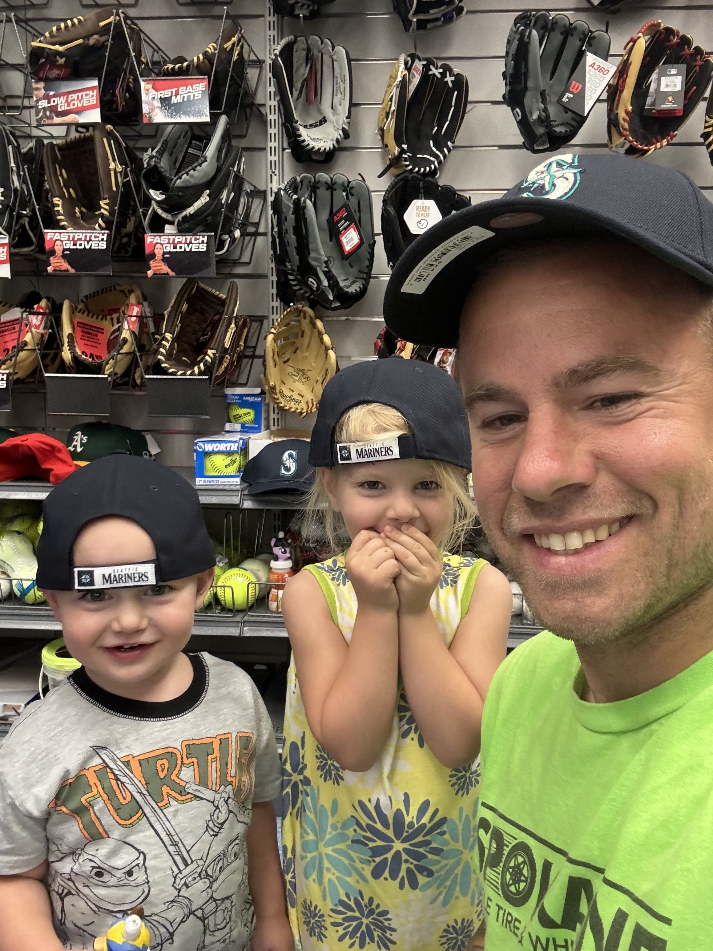 Two children and an adult taking a selfie in sports equipment store, with baseball gloves, caps, and sports supplies on the shelves behind them.
