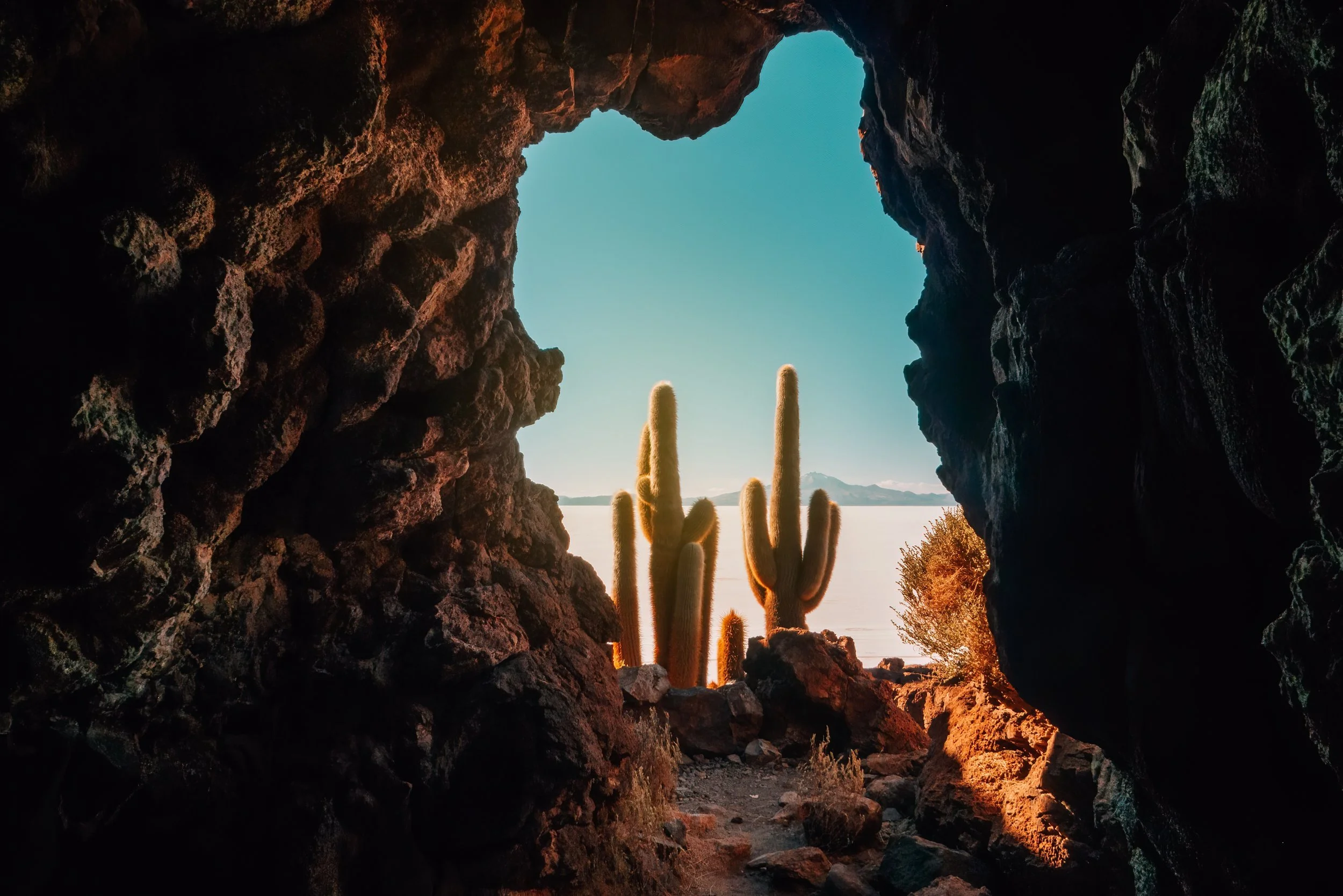 View through a rocky cave opening revealing desert landscape, tall cacti, and mountains under a clear blue sky.