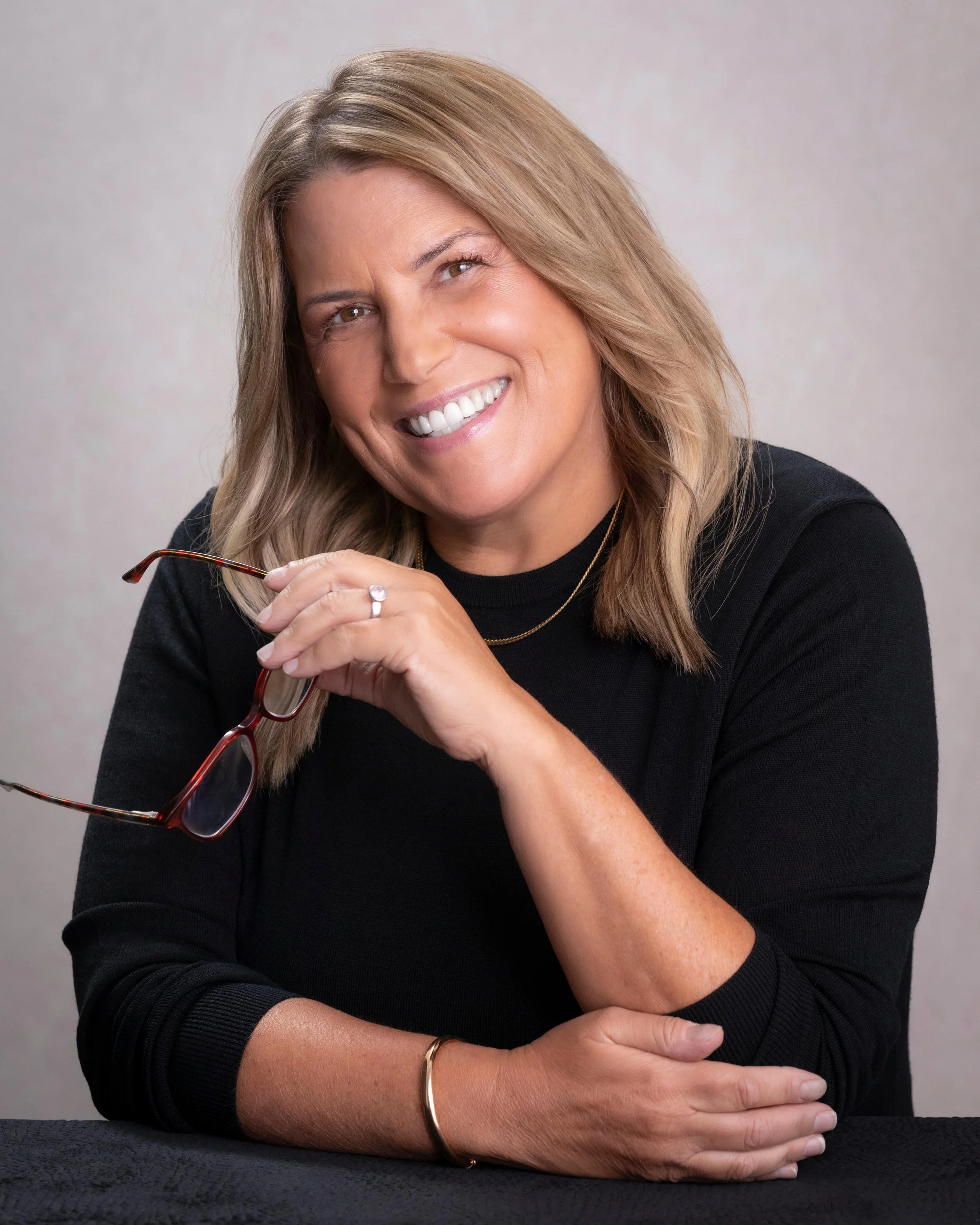 A woman smiling, holding glasses in her hand, sitting at a table with a grey background.