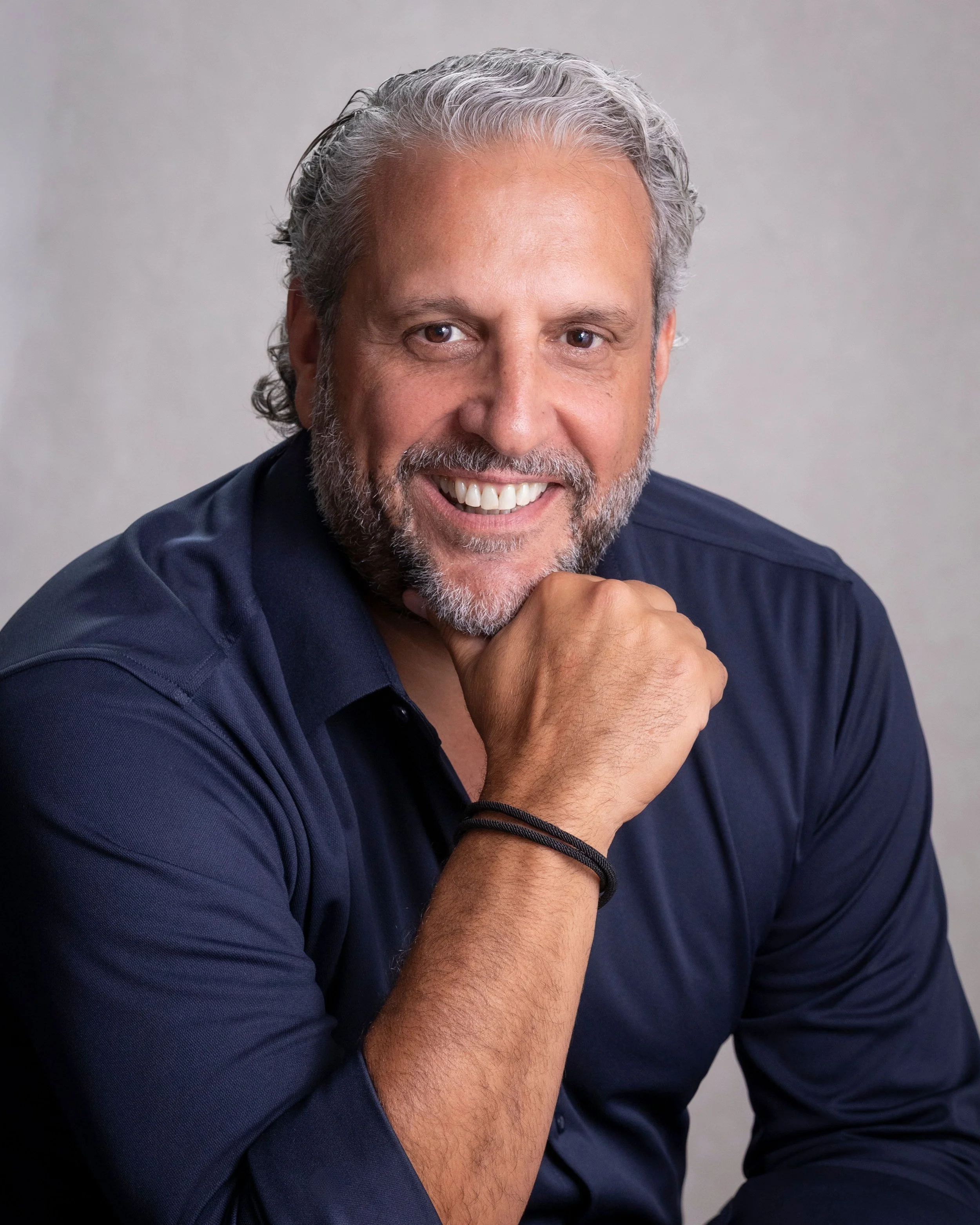 A smiling man, with his hand under his chin against a plain gray background.
