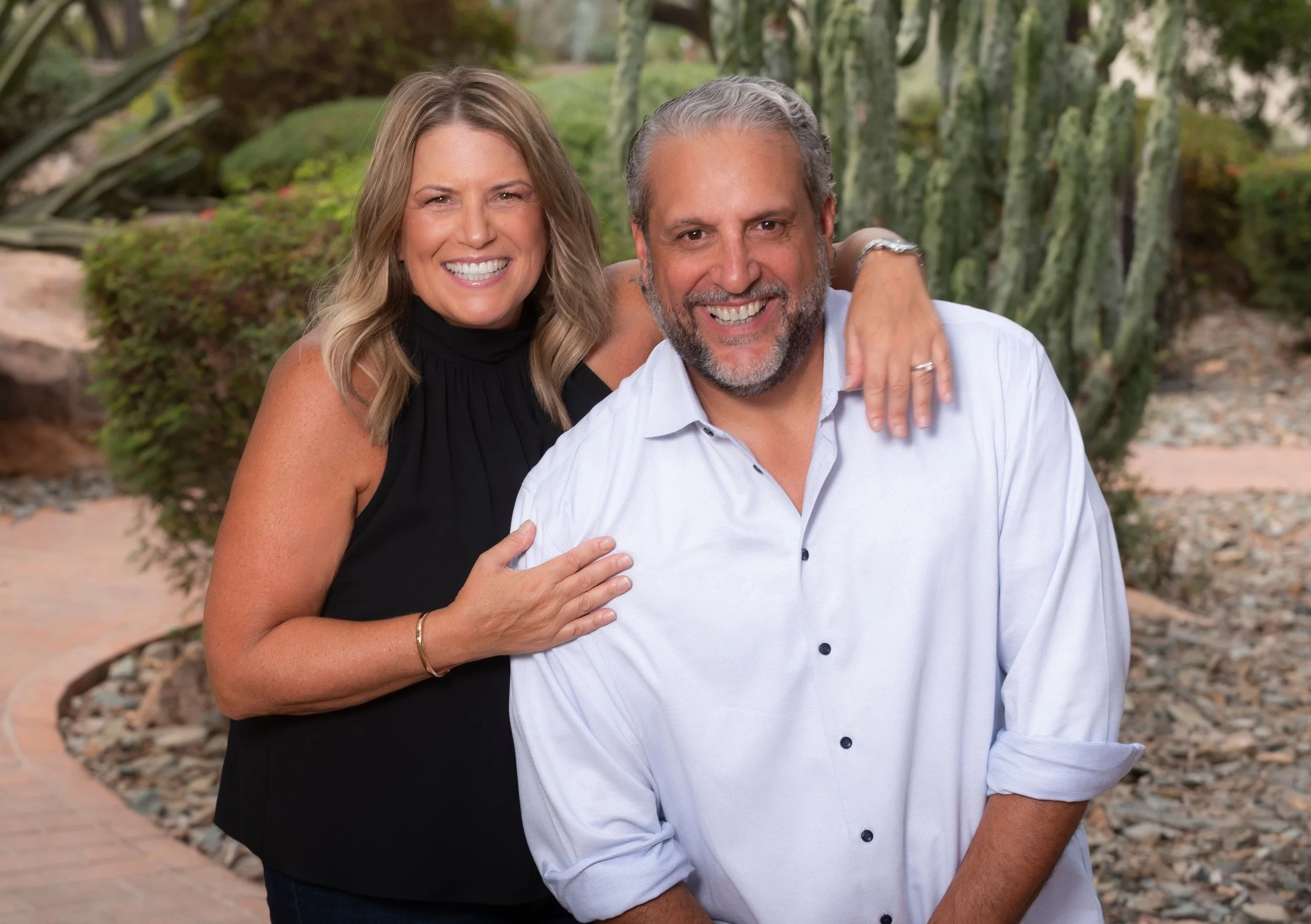 A smiling woman standing behind a man, outdoors with greenery