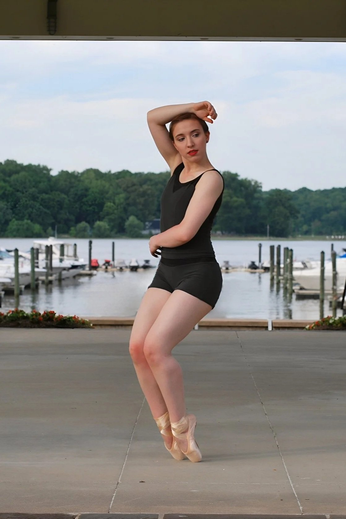 A young woman practicing ballet outdoors near a body of water with boats, wearing a black tank top and shorts, en pointe.