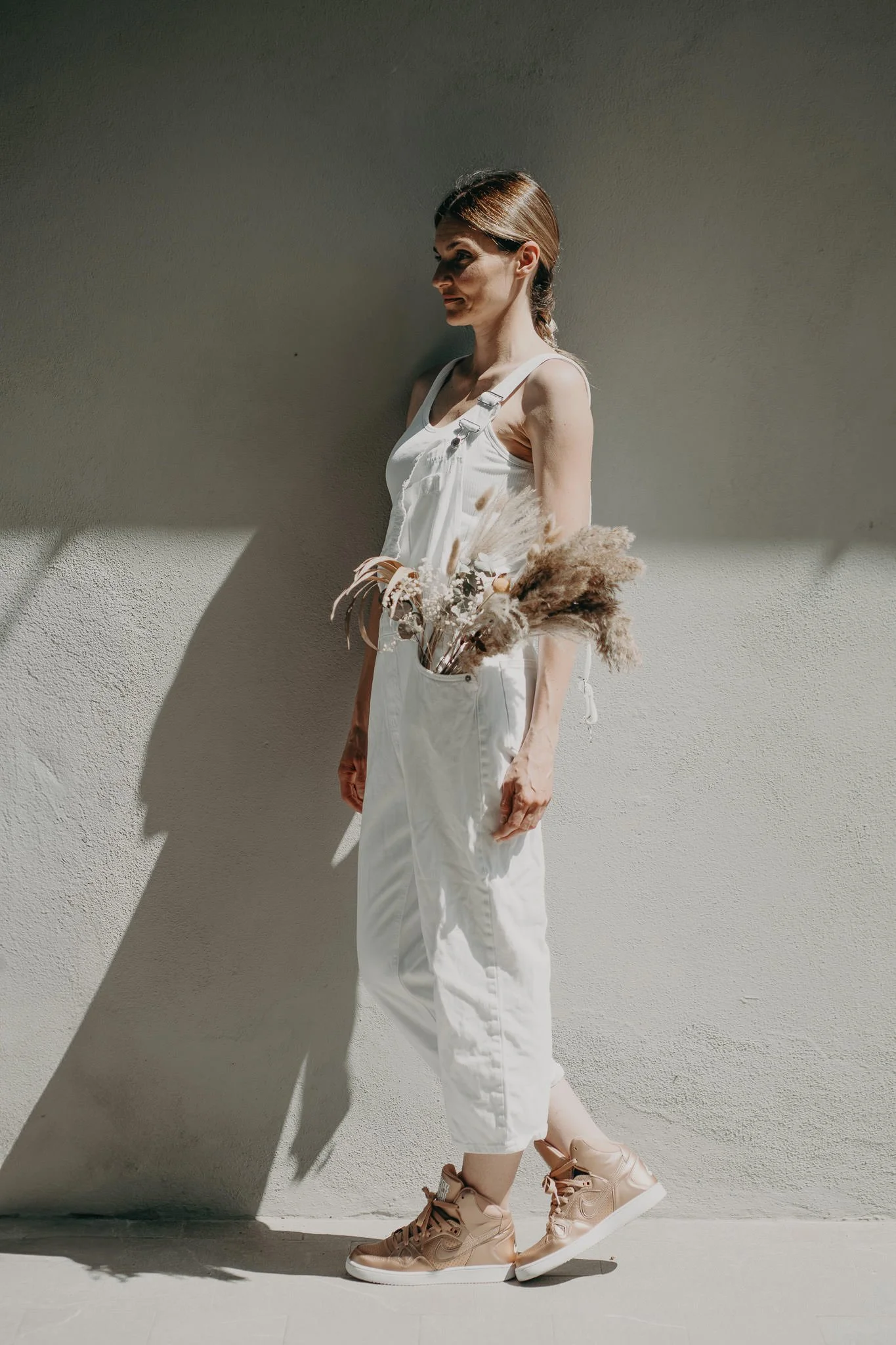 Donna giovane con lacci ai piedi, pantaloni bianchi, maglietta bianca e abiti beige, che tiene un mazzo di fiori secchi, in piedi contro un muro chiaro con luce solare che crea un'ombra.